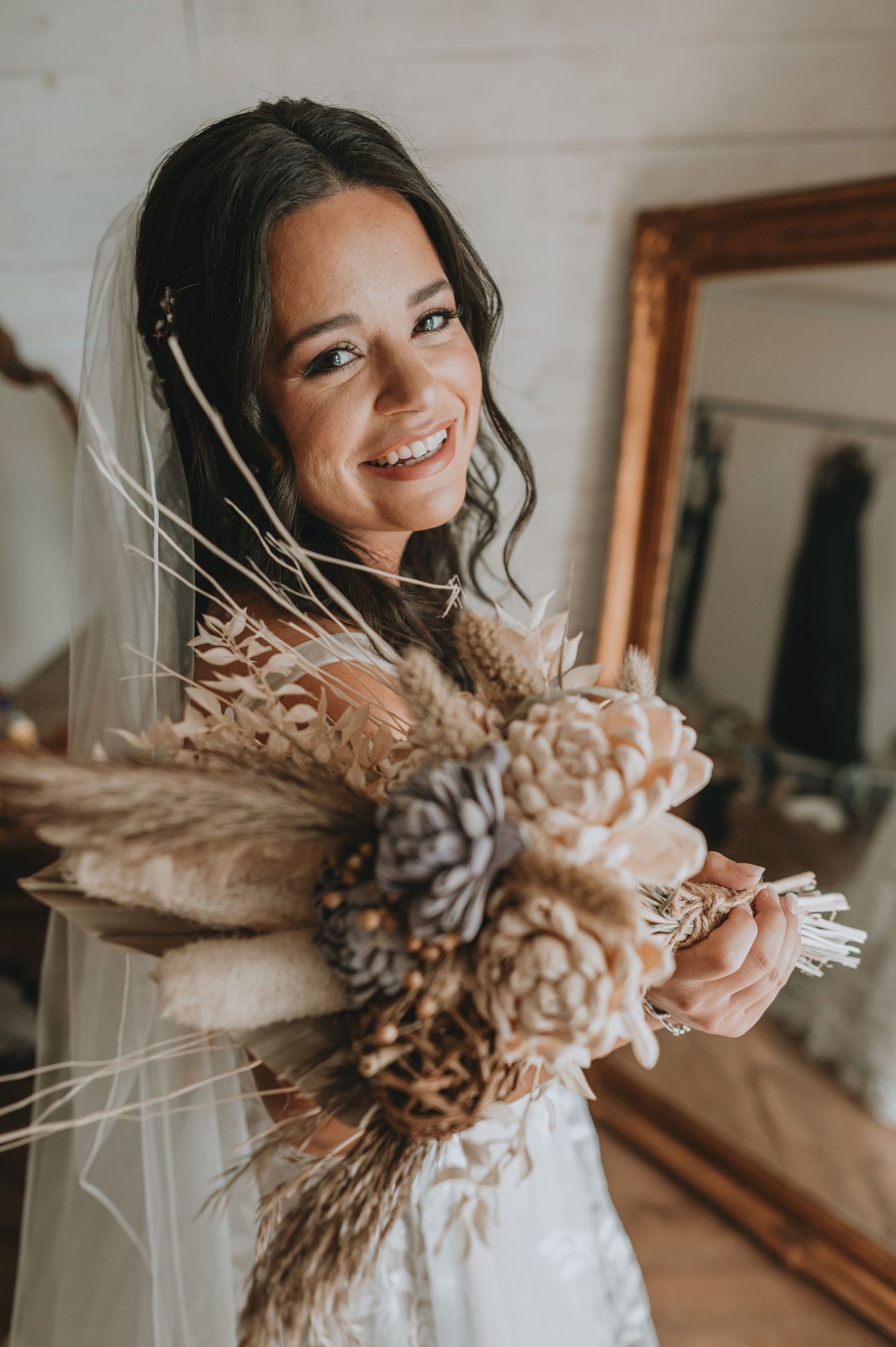 Smiling bride in white veil holding a boho dried flower bouquet with pinecones and pampas grass indoors.