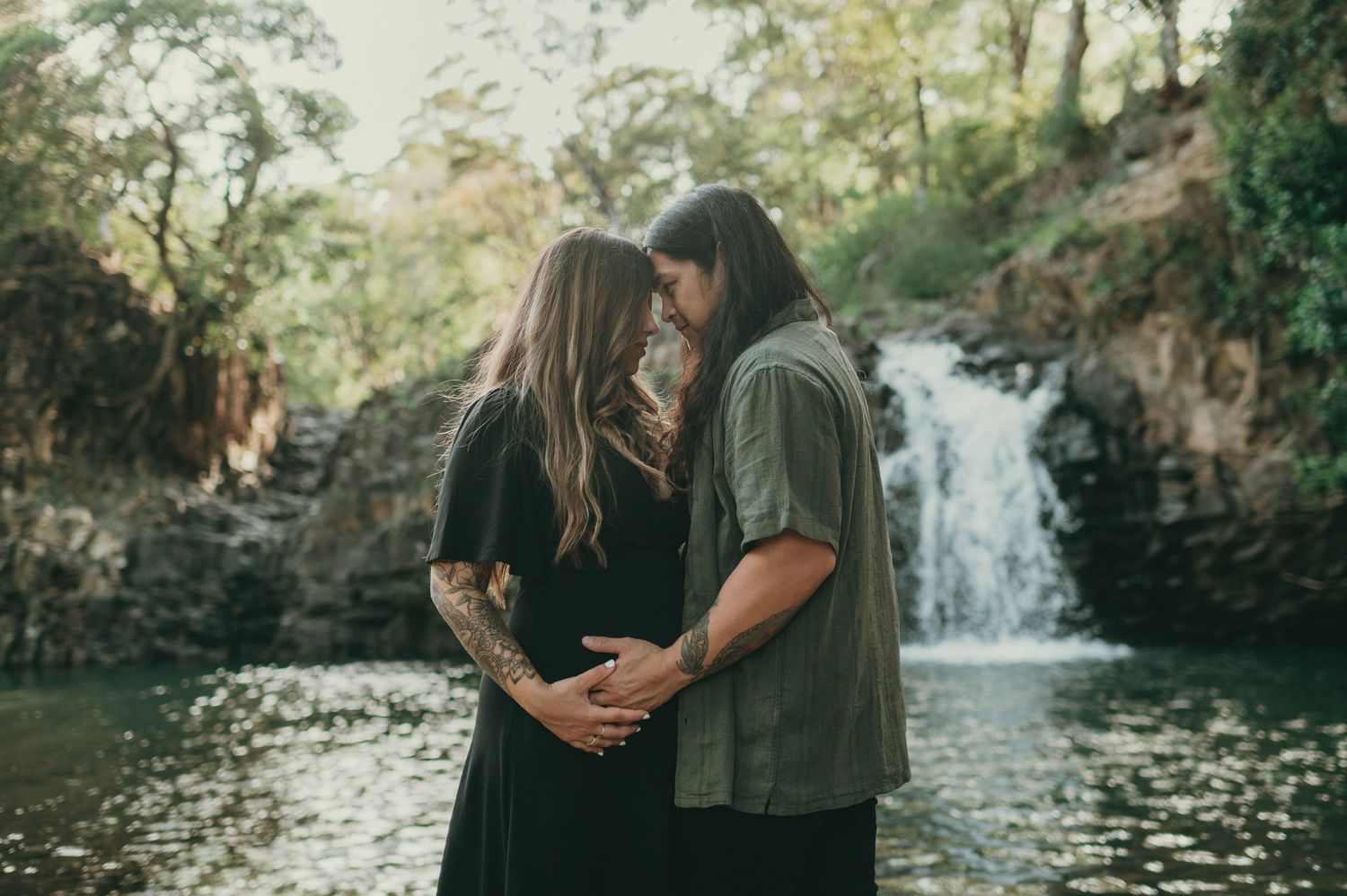 Couple embracing tenderly in front of a serene waterfall surrounded by lush greenery, maternity photo session outdoors.