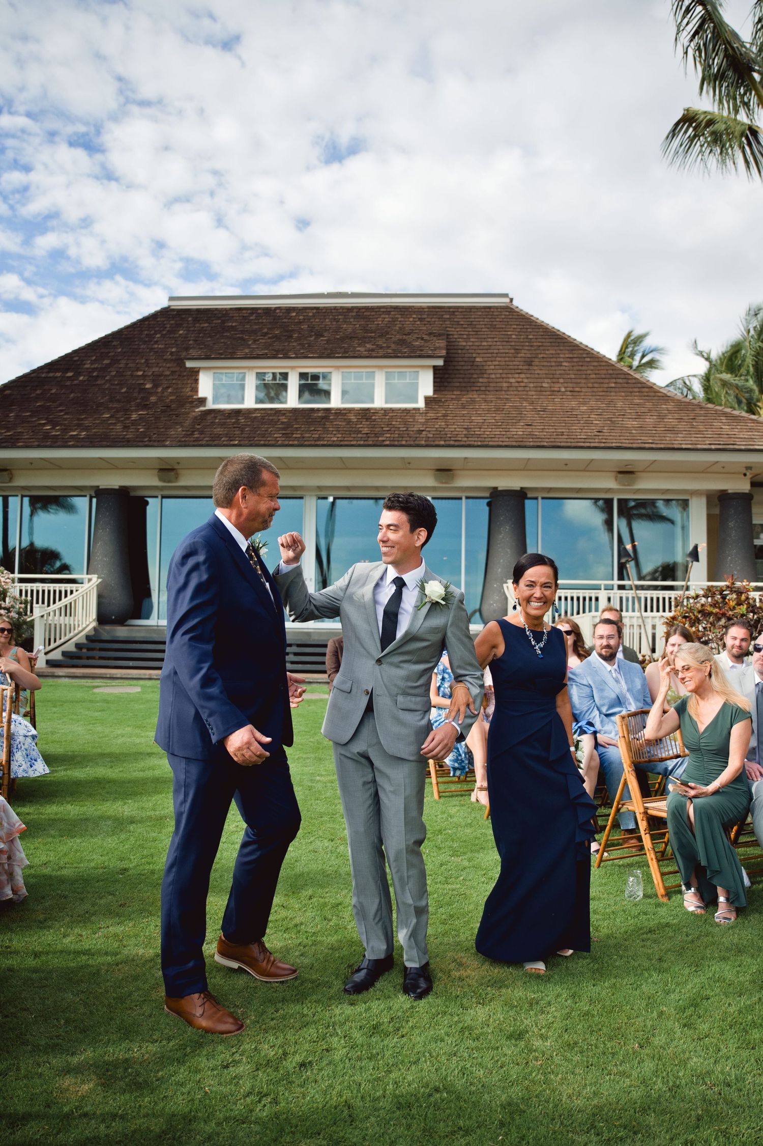 Groom walks down outdoor wedding aisle escorted by parents on a sunny lawn with guests seated nearby.