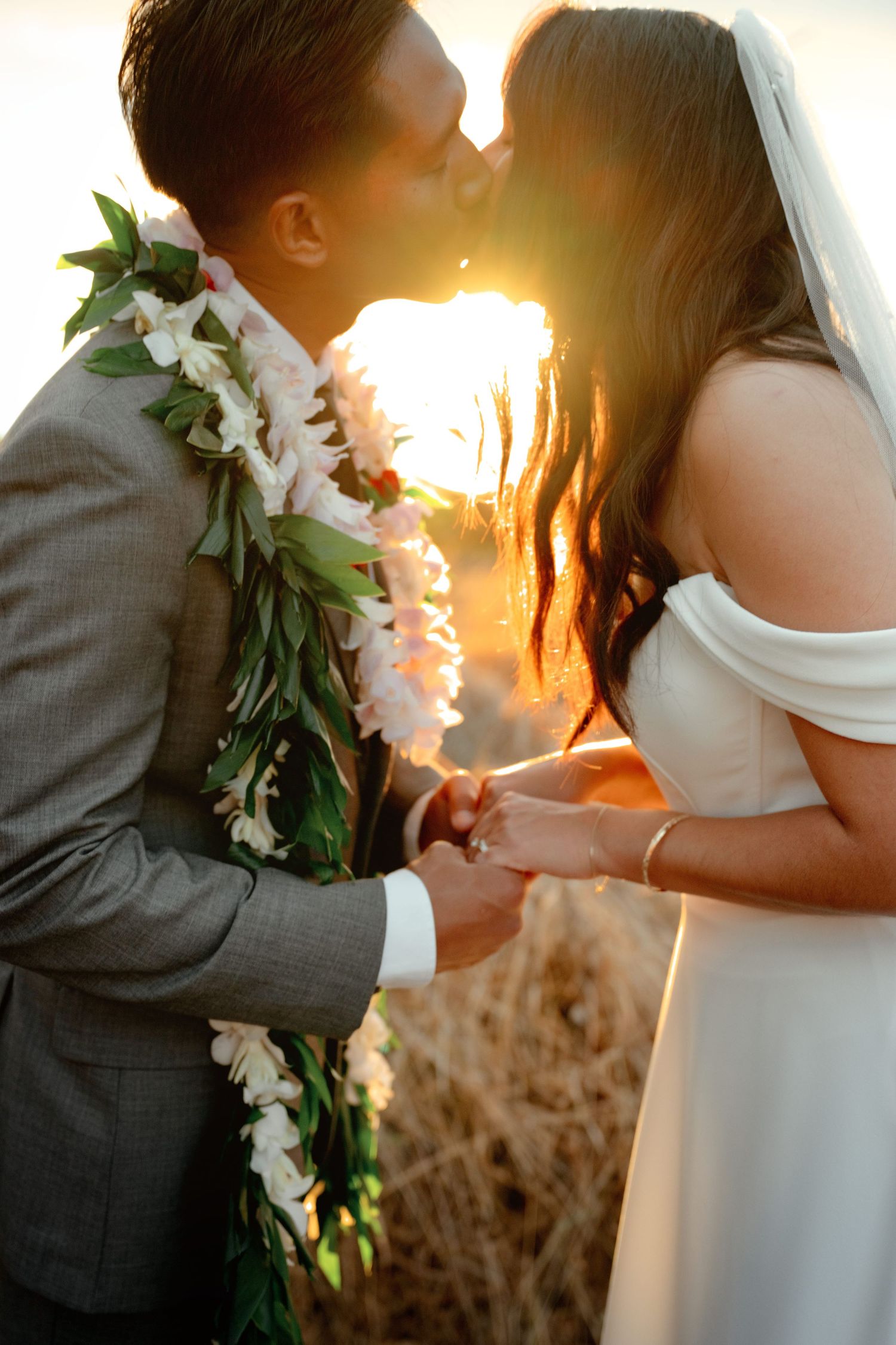 Bride and groom kissing at golden sunset, groom wearing floral lei, bride in white off-shoulder gown with veil.