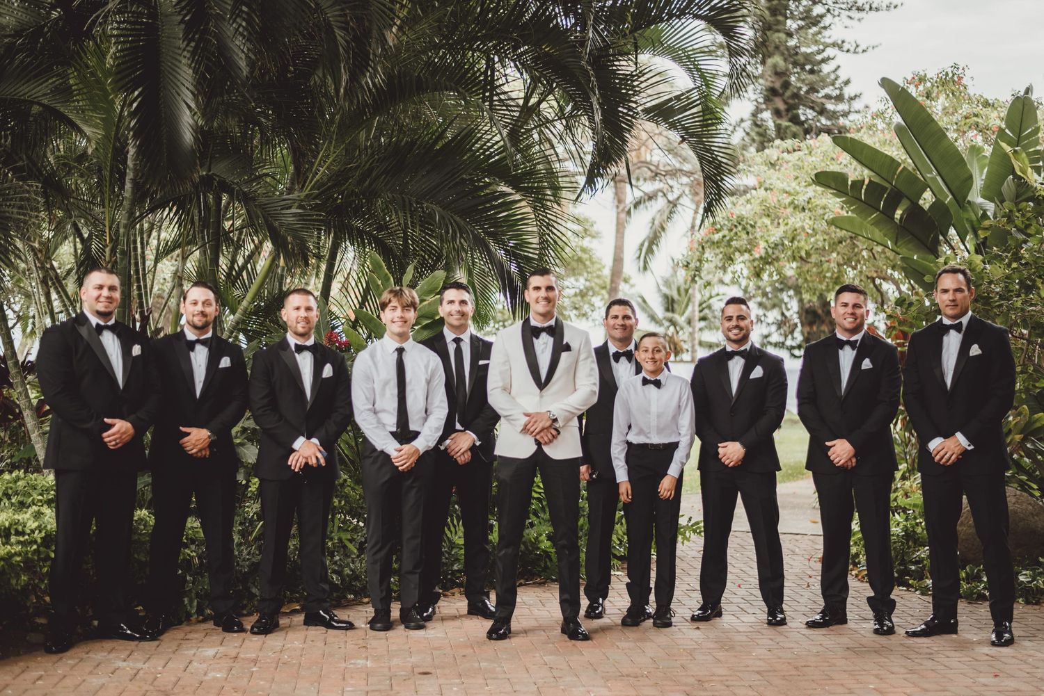 Groom in white tuxedo jacket stands with groomsmen in black tuxedos among lush tropical palm trees.