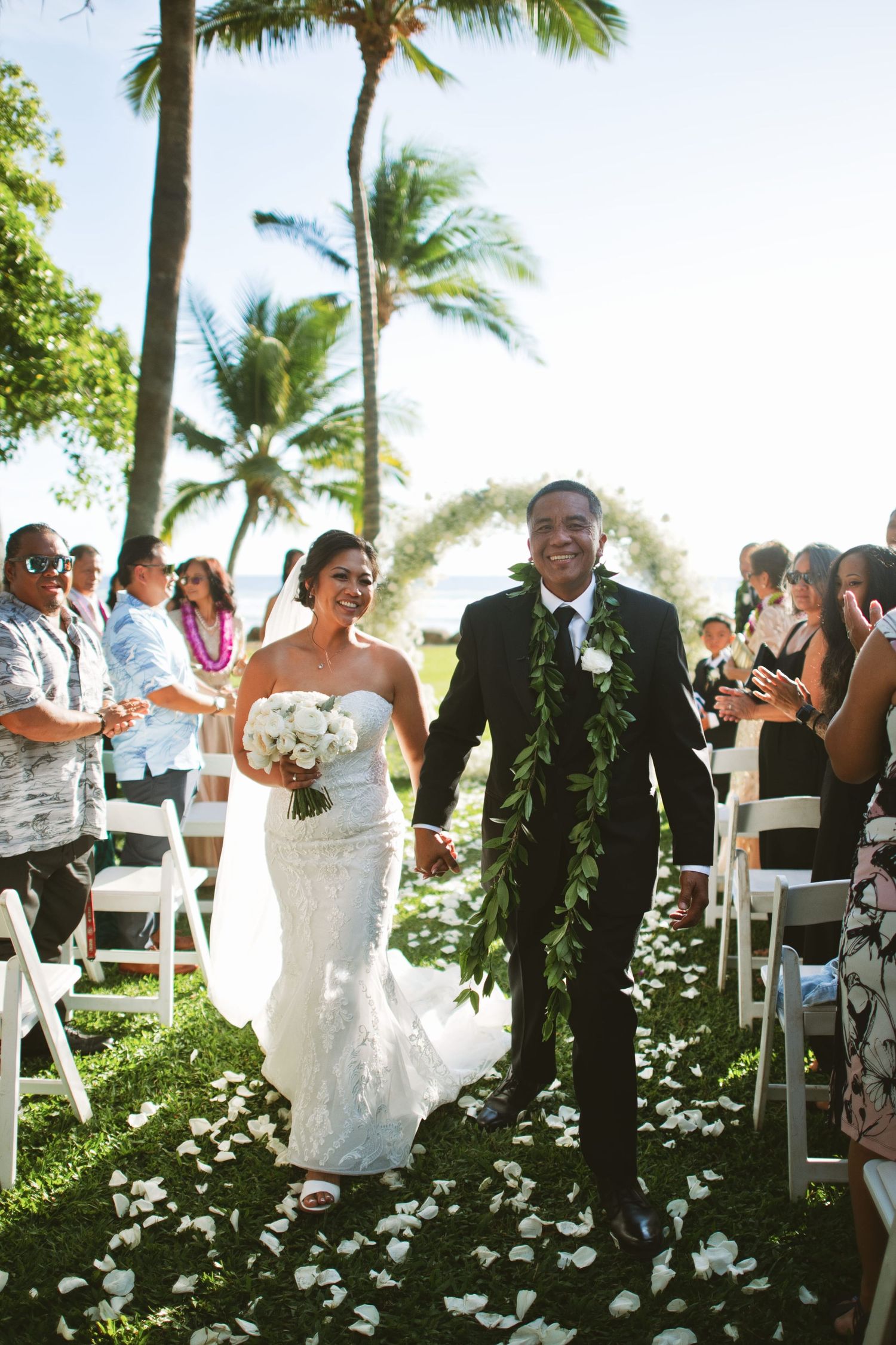 Newlywed couple walks down petal-lined aisle at outdoor Hawaiian wedding ceremony surrounded by palm trees and cheering guests.