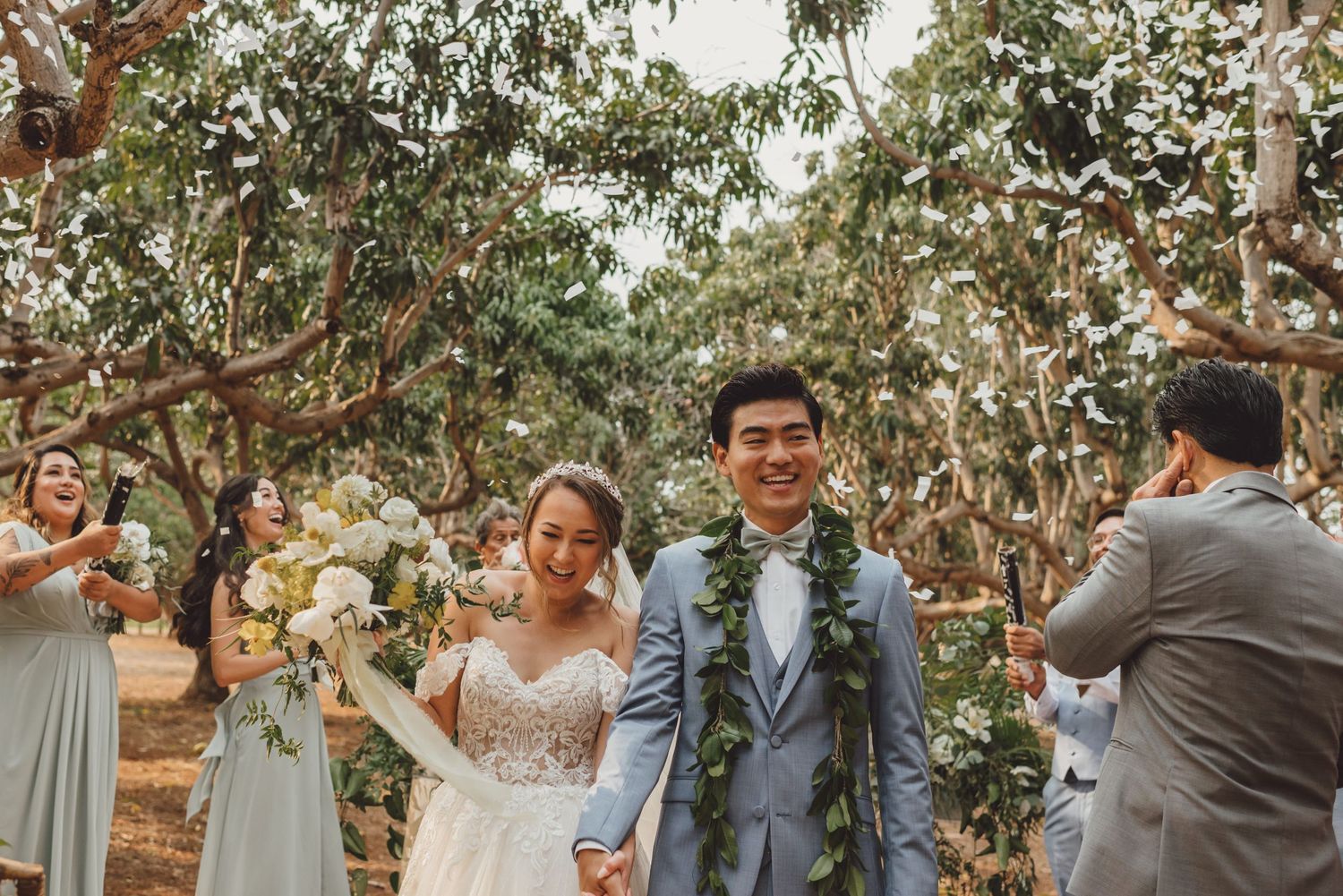 A joyful Asian couple walks hand-in-hand at an outdoor wedding ceremony as guests in sage green attire toss confetti.