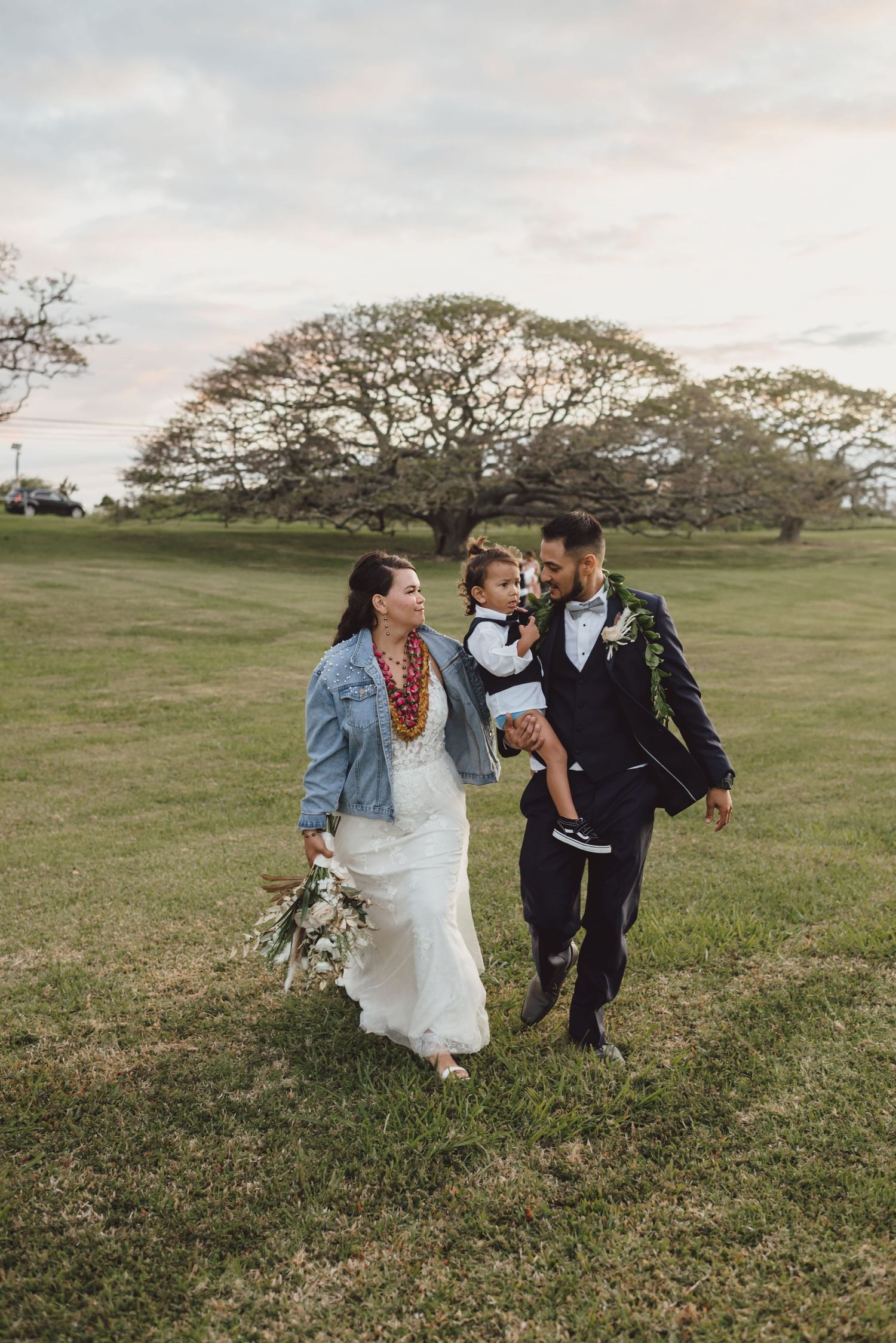 Bride and groom walking on green grass with child, large tree backdrop, wedding bouquet, denim jacket, floral lei.
