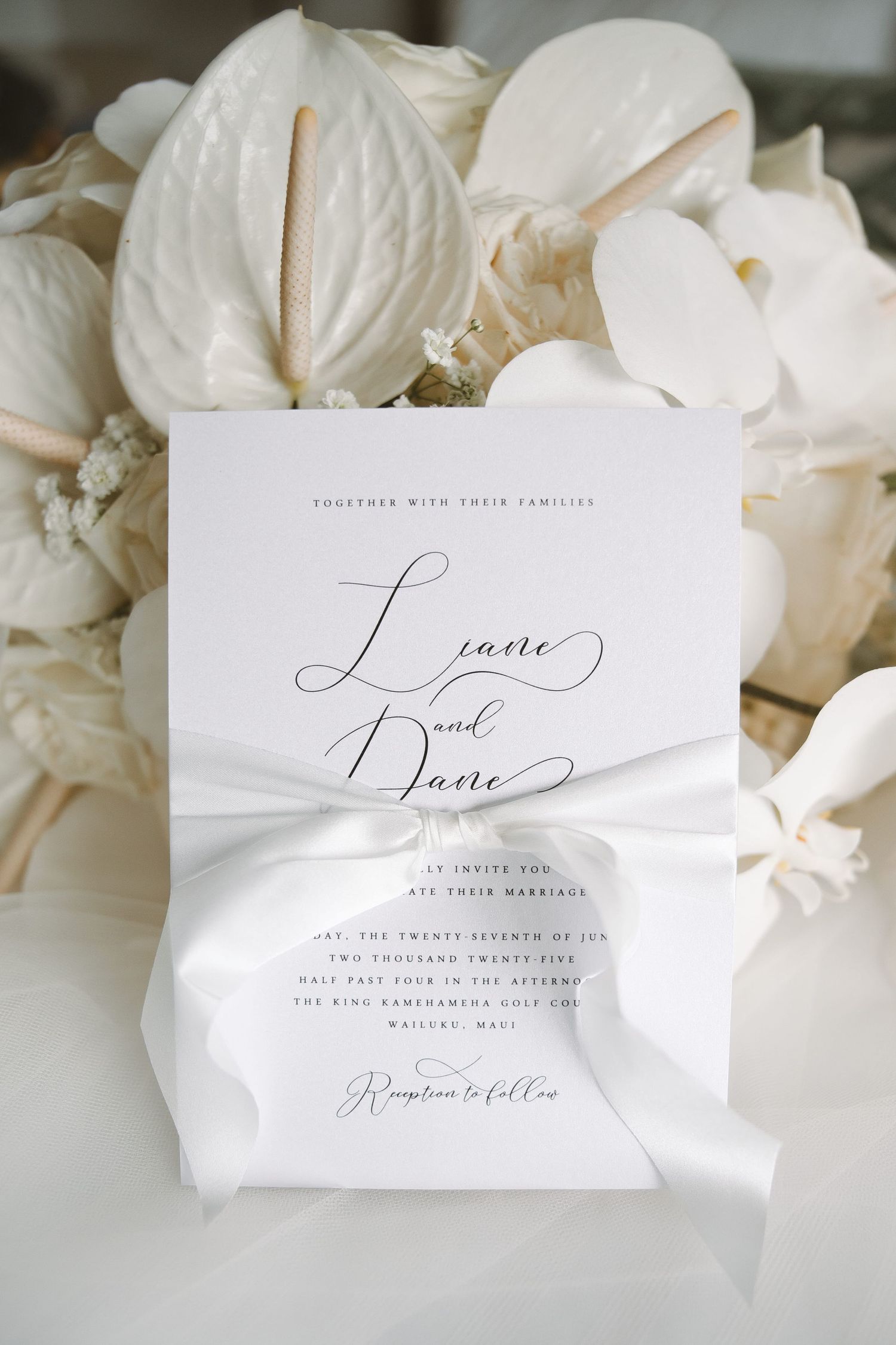 Elegant white wedding invitation with calligraphy script tied with satin ribbon, displayed against white floral bouquet.