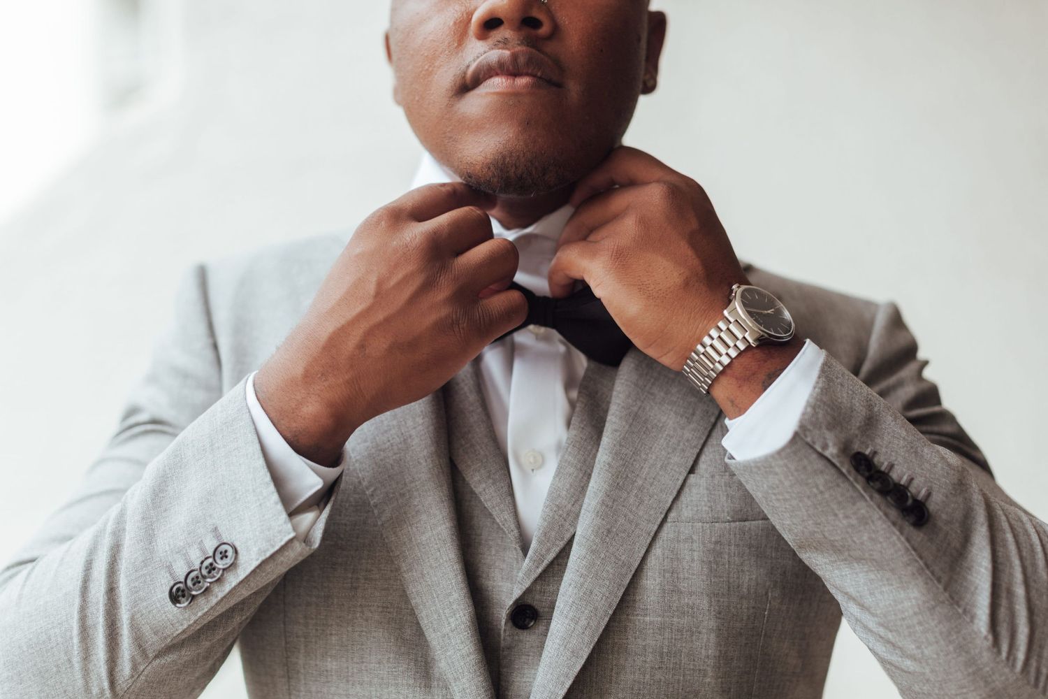 Man in gray suit adjusting black bow tie, wearing silver watch, dressed elegantly for formal occasion.