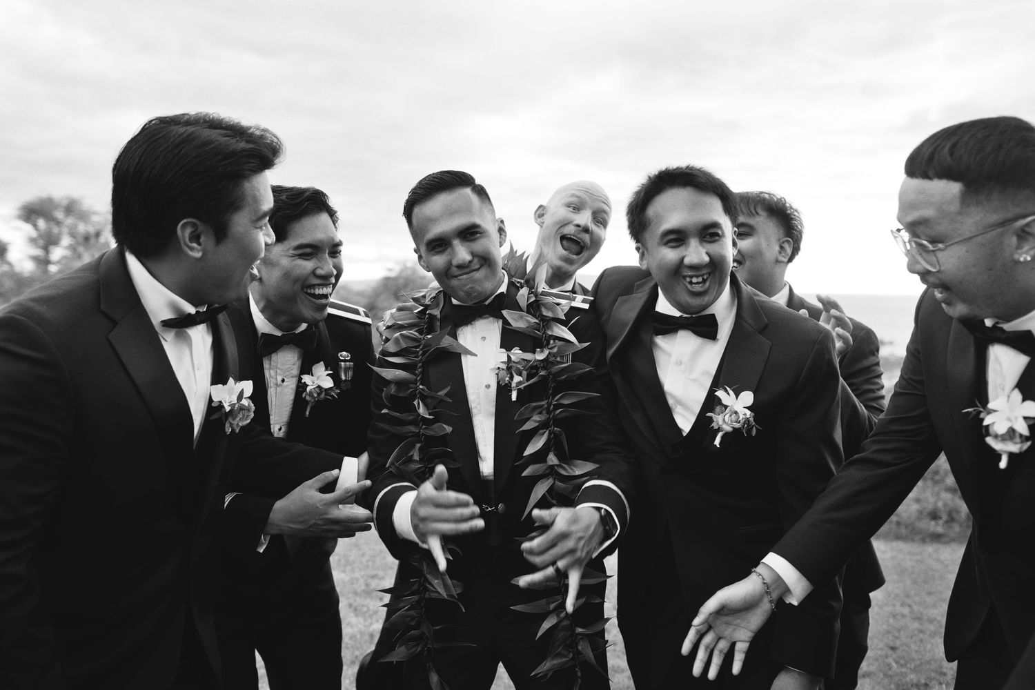 Black and white photo of groom laughing with groomsmen in tuxedos outdoors at a wedding celebration.