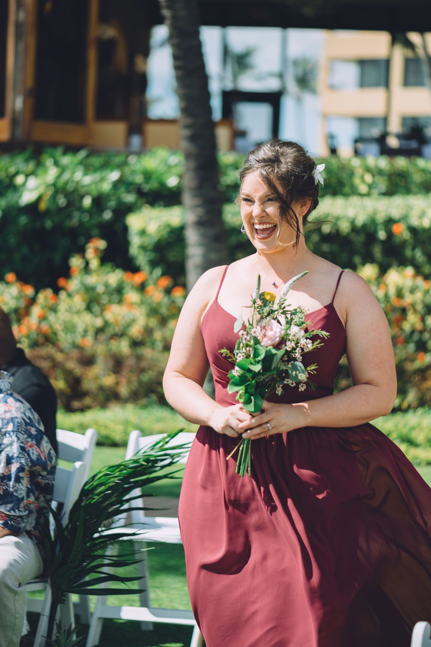 Bridesmaid in burgundy dress holds floral bouquet at outdoor tropical wedding venue.