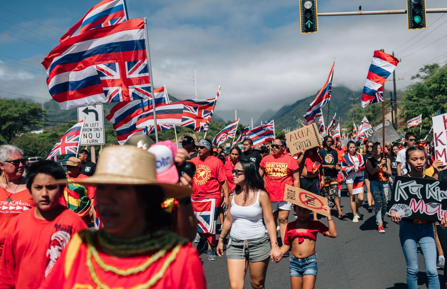 Hawaiian flag bearers lead protest march through intersection with mountains in background.