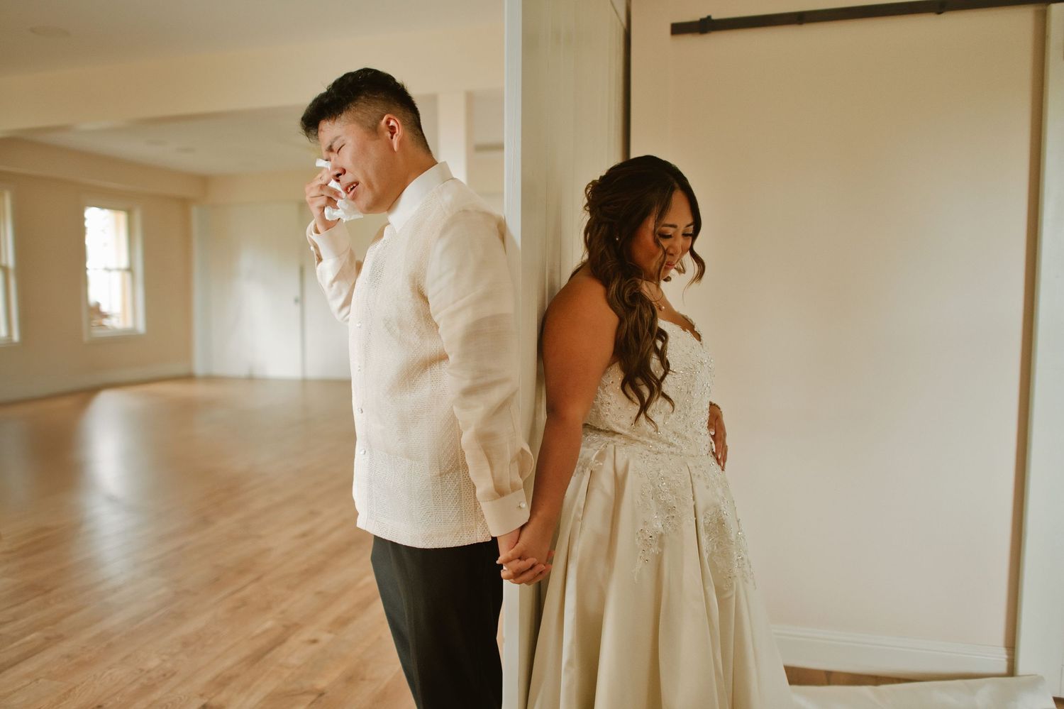 Bride and groom share an emotional first look moment, holding hands back-to-back in a bright, airy wedding venue.