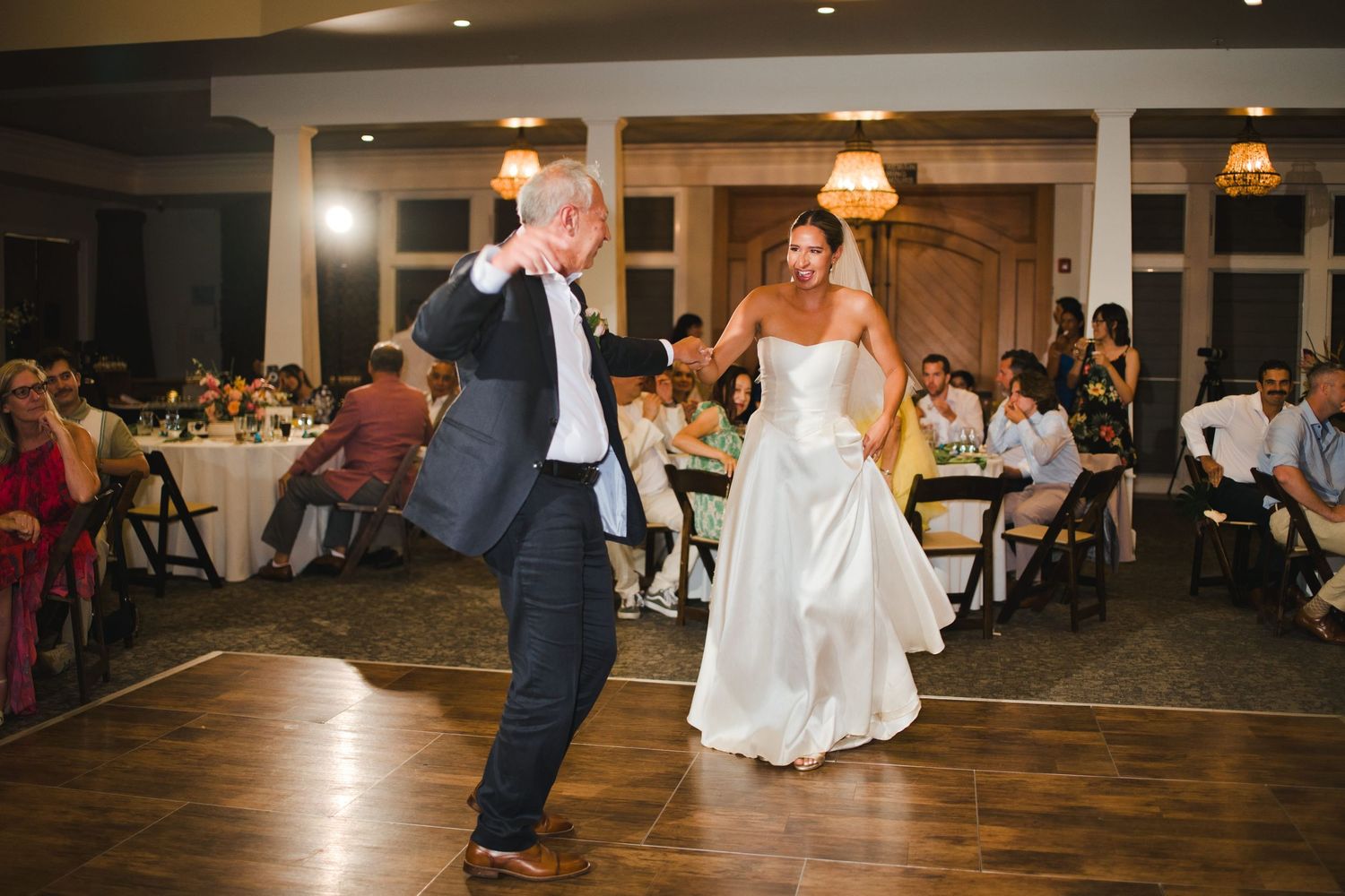 Bride and groom share a lively first dance at elegant wedding reception with guests seated in background.