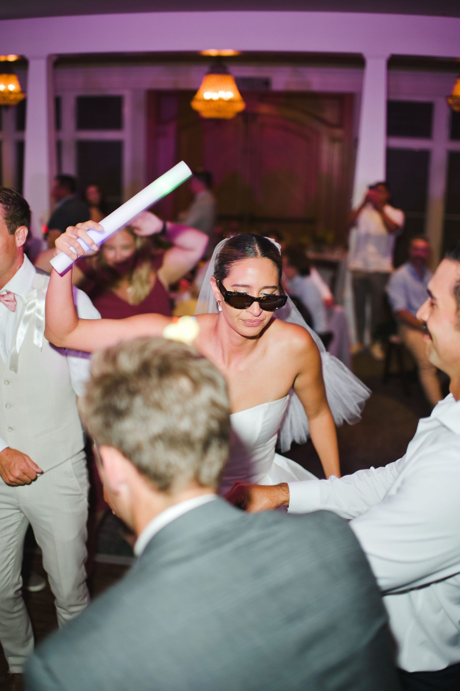 Bride in white dress and sunglasses dancing with glow stick at lively wedding reception party.