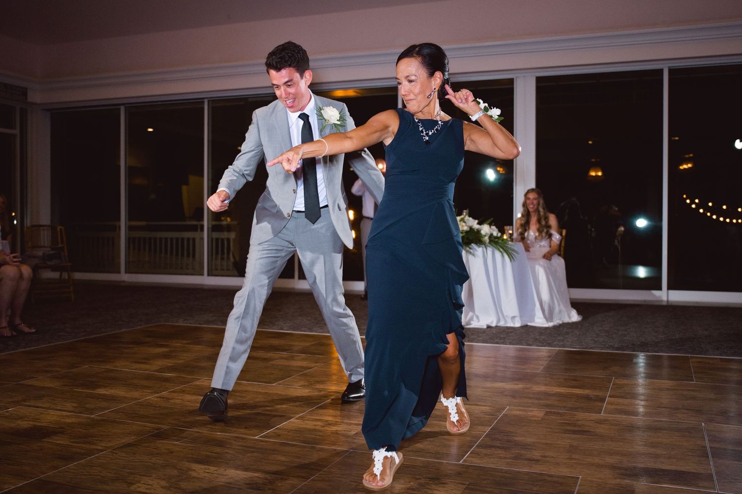 Groom in gray suit and woman in navy dress dancing energetically at wedding reception on dance floor.