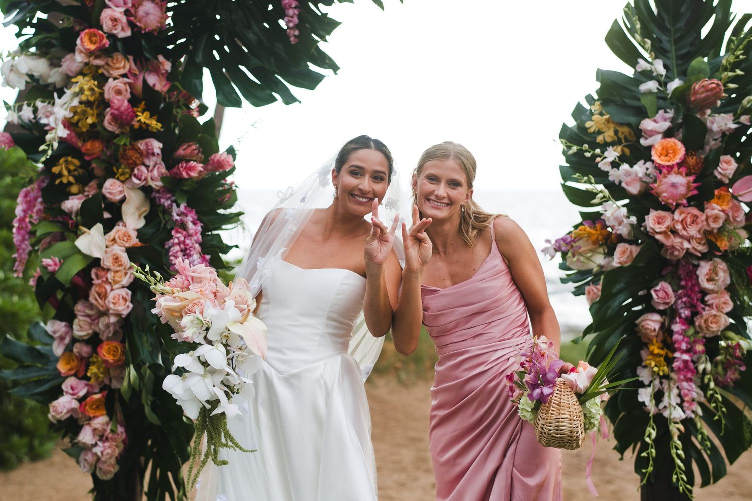 Bride in white gown and bridesmaid in pink dress smiling under floral arch with tropical flowers at beach wedding.