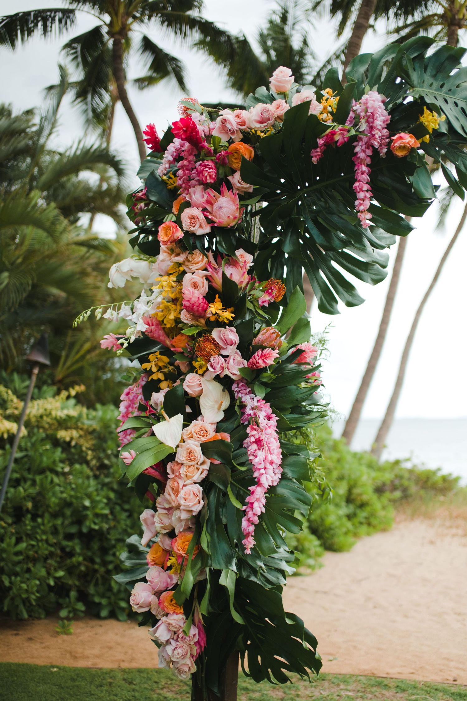 Tropical wedding arch adorned with vibrant pink, orange, and white flowers with lush monstera leaves on a beachside setting.
