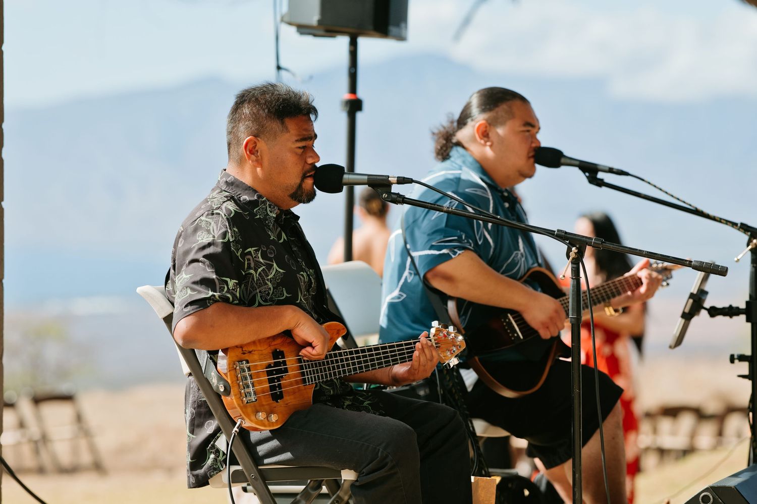 Two musicians playing guitar and bass at an outdoor event with mountains in the background.