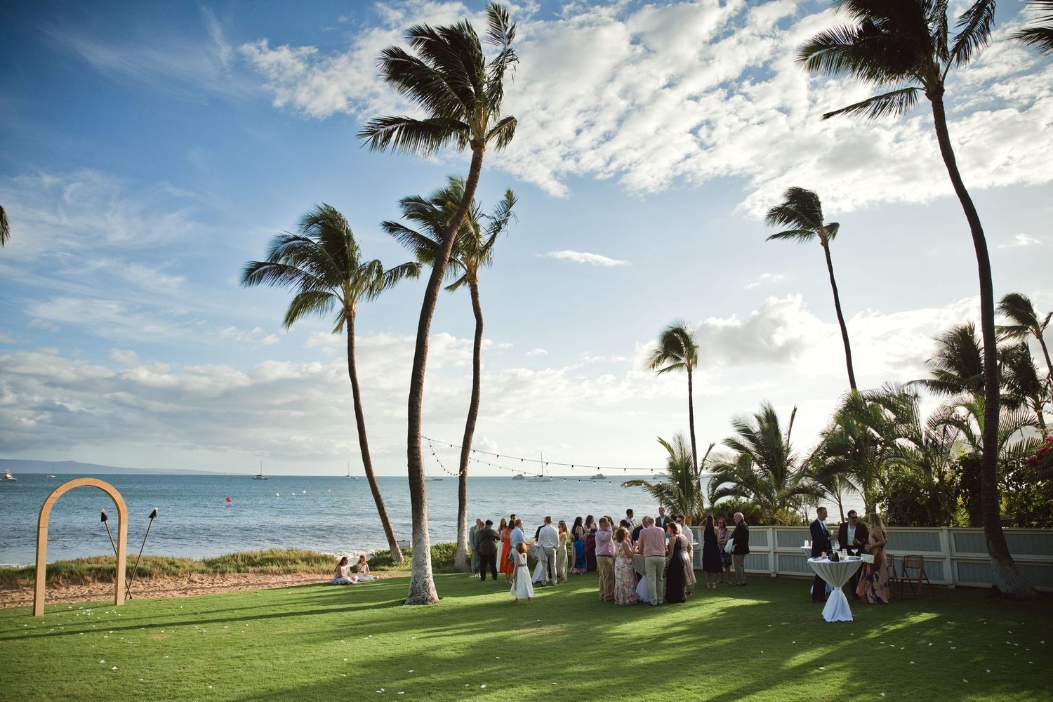 Outdoor beach wedding ceremony on lush green lawn with tall palm trees and ocean views in Hawaii.