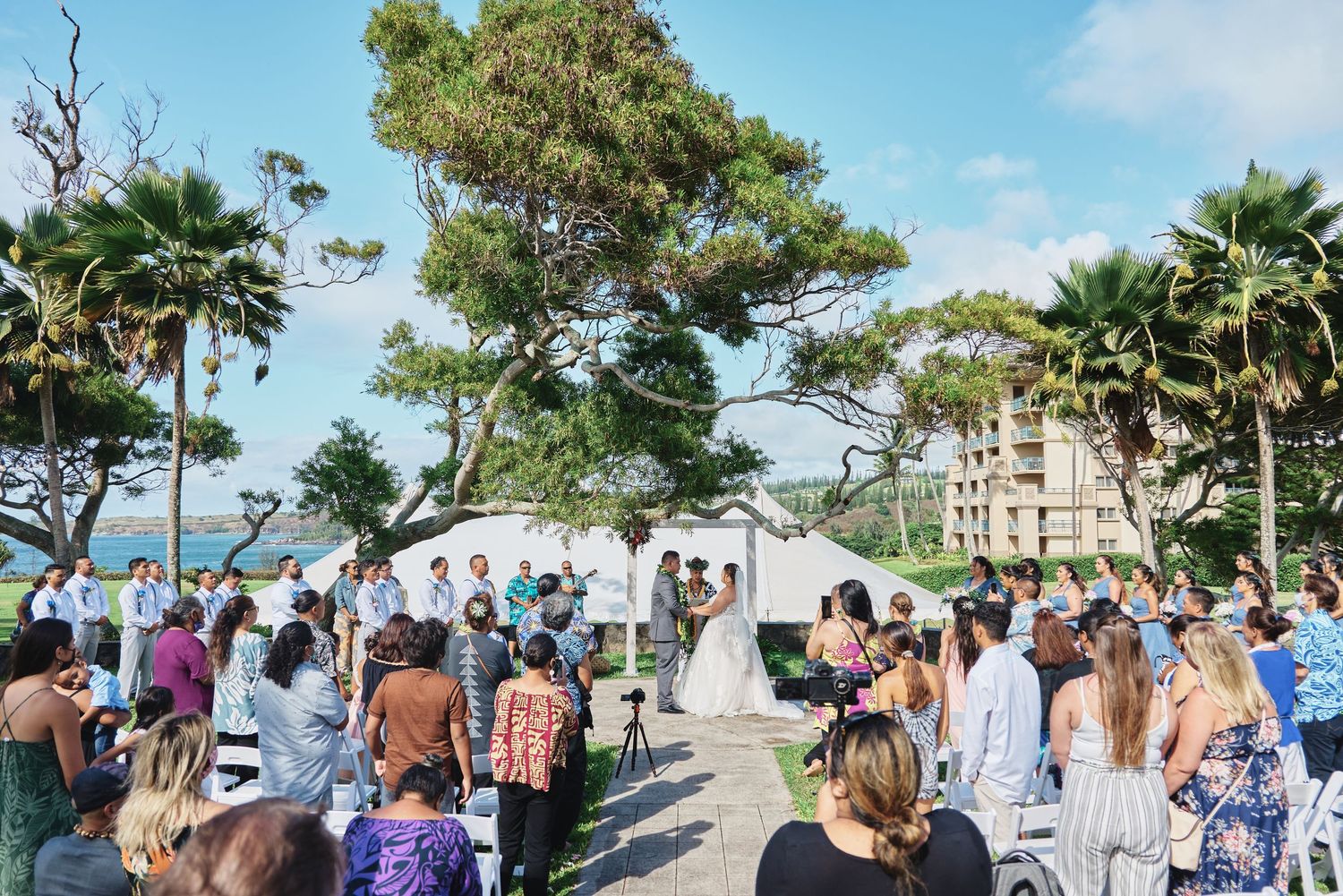Guests gather for outdoor beachside wedding ceremony under blue skies.