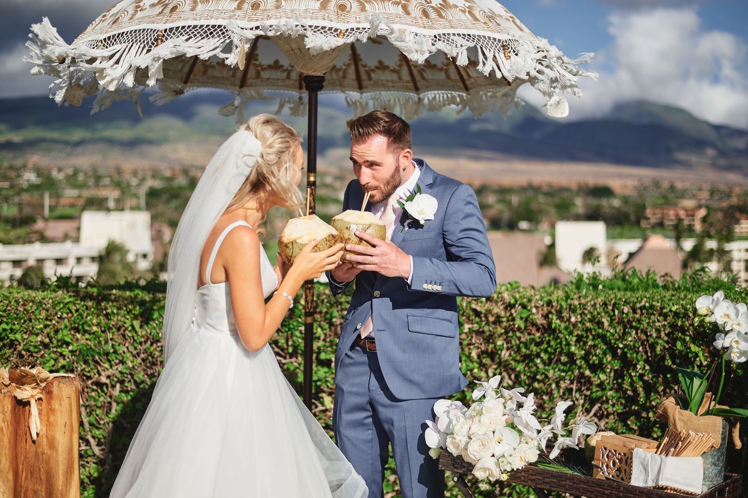 Newlyweds share a drink under a rustic umbrella at an outdoor vineyard wedding with mountains in the background.