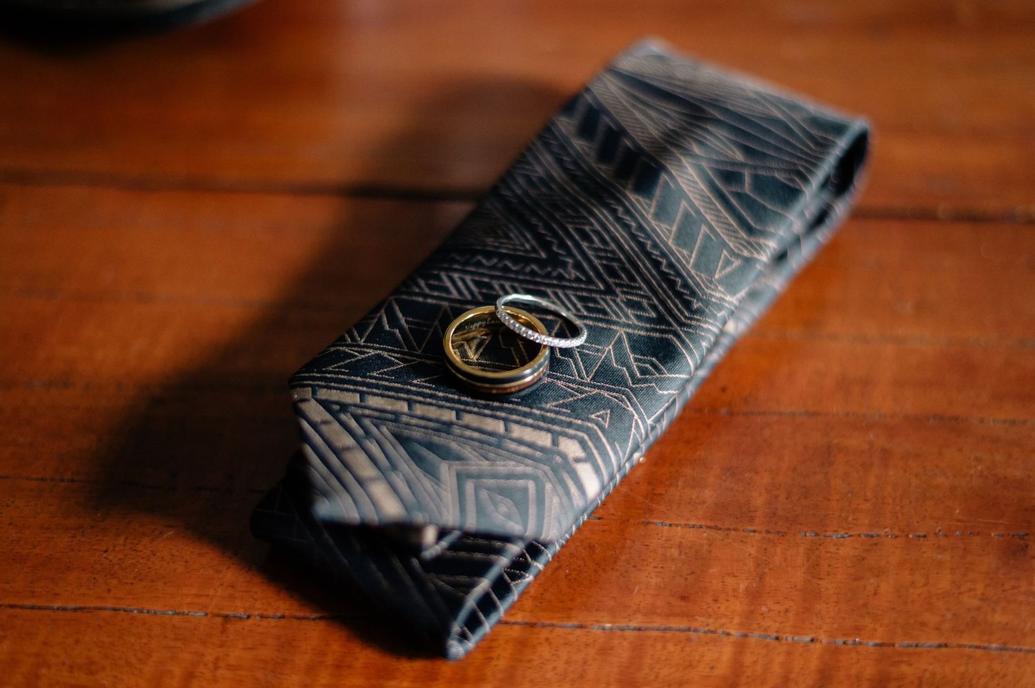 Wedding rings rest on a patterned navy blue tie on wooden surface.