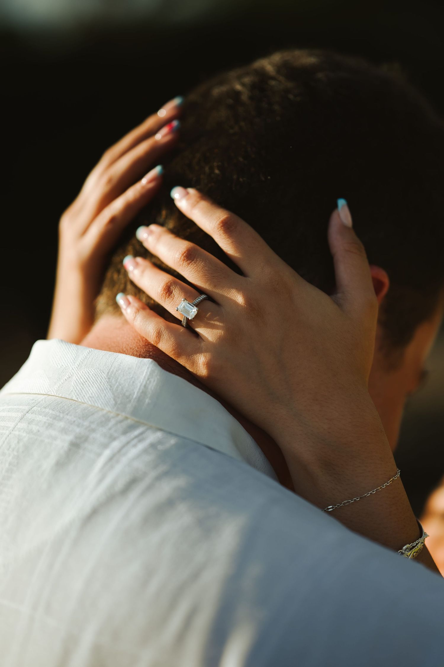Close-up of womans hands with diamond engagement ring and blue nails on mans neck.