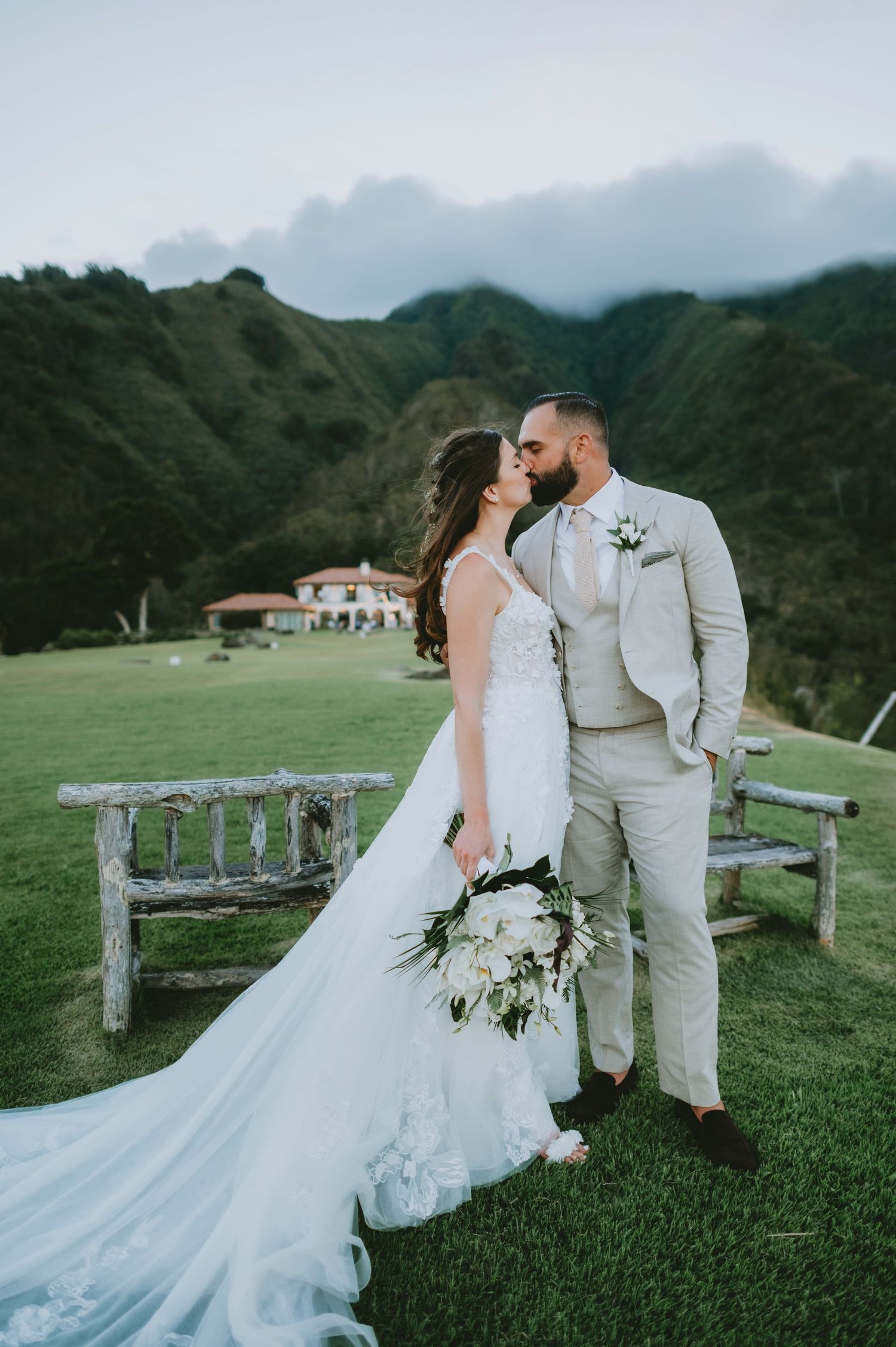 Bride and groom share a romantic kiss on lush green hillside venue with mountain backdrop on their wedding day.