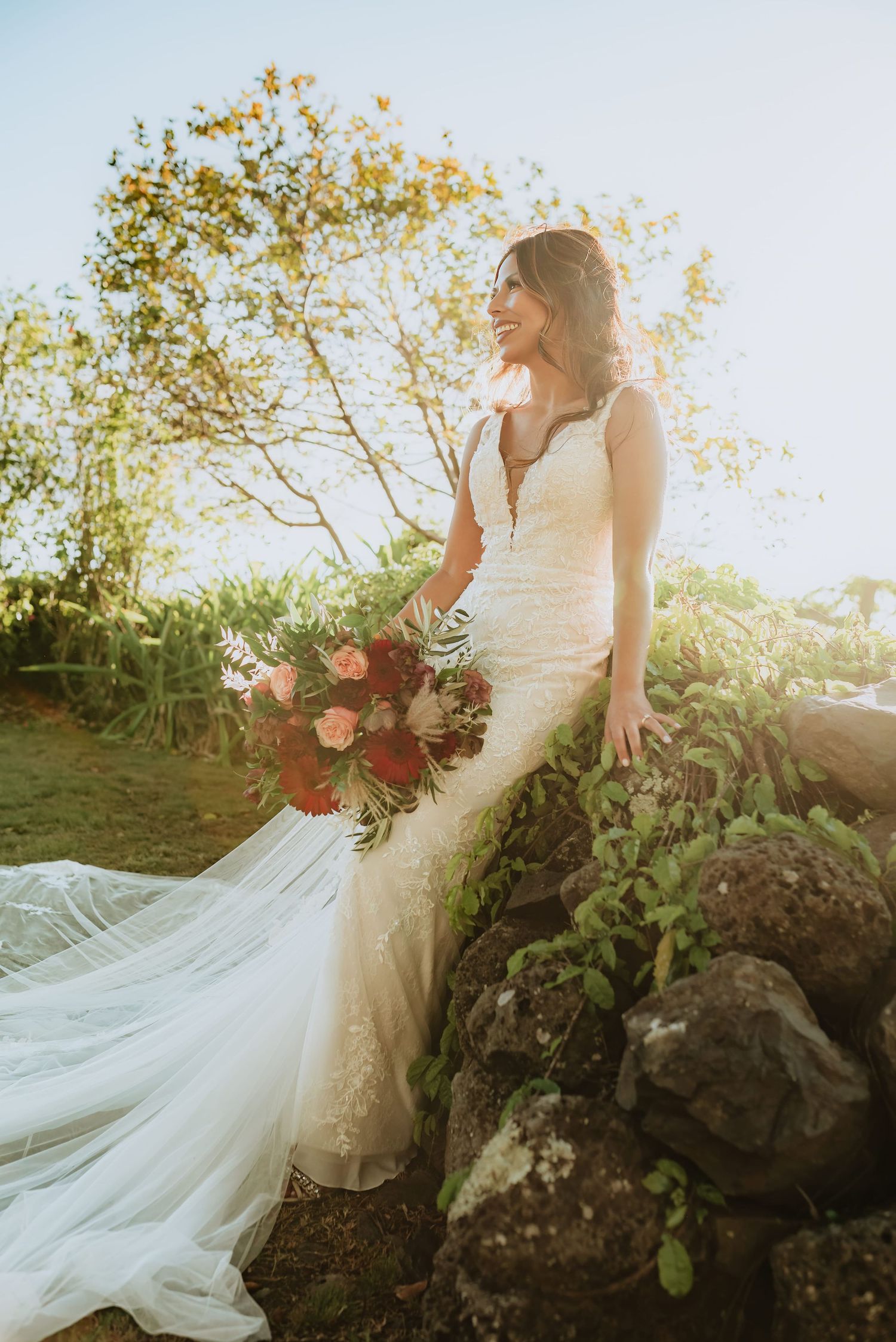 Bride in lace mermaid gown holding red bouquet poses on rocky garden wall with sunlit trees in background.