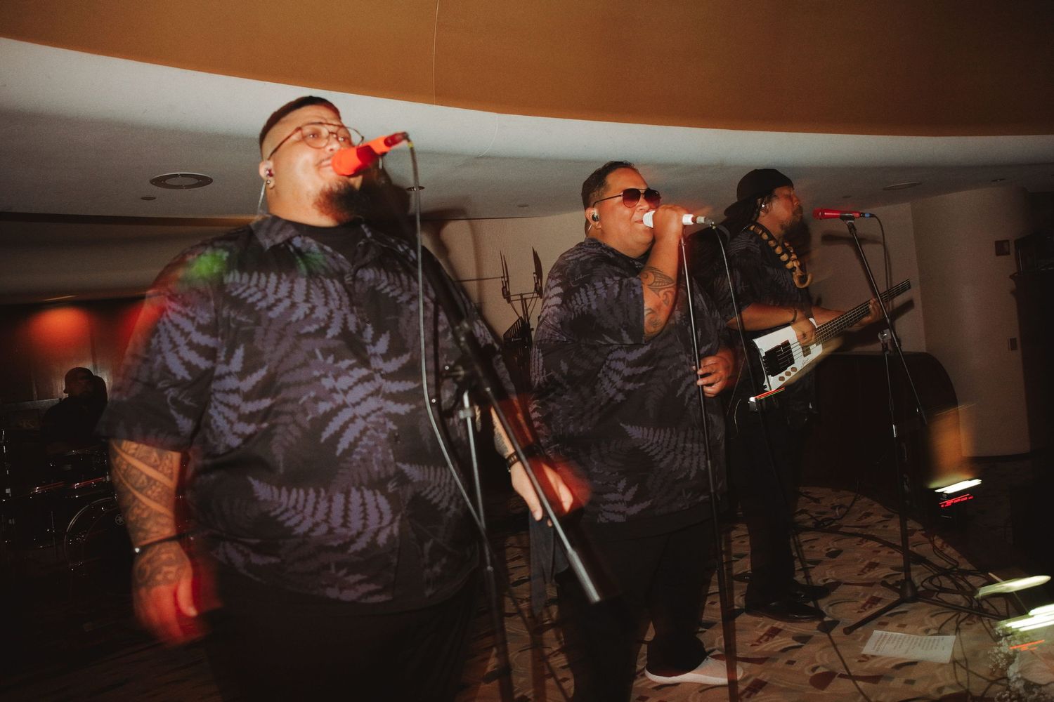 Three musicians in matching patterned shirts perform on stage with microphones and instruments at an indoor venue.