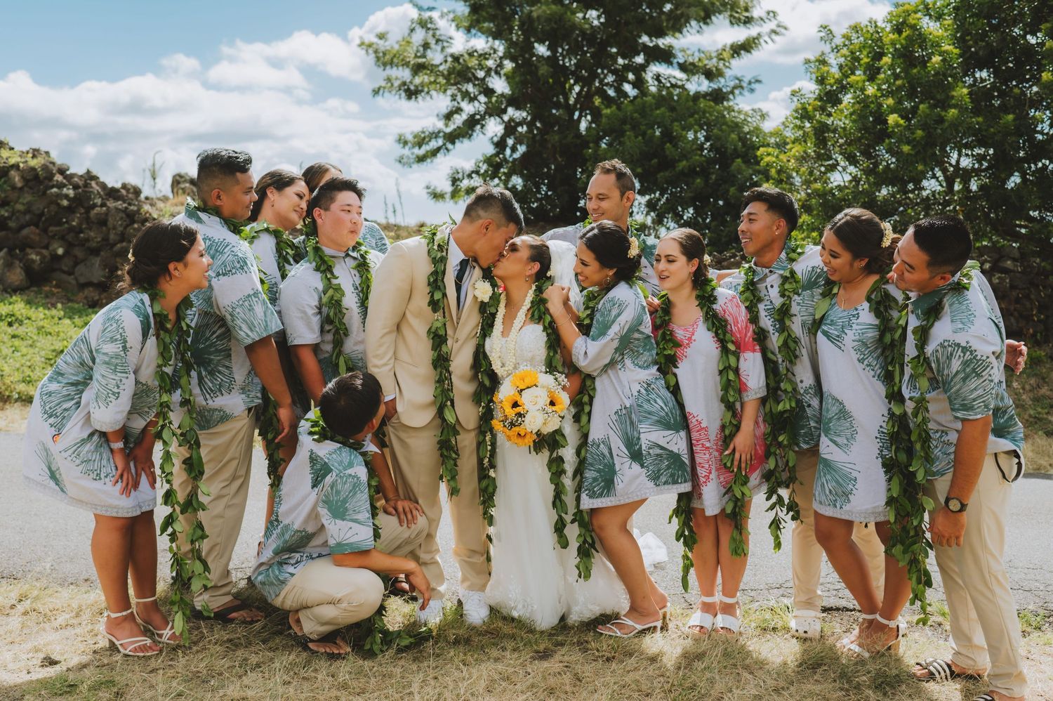Wedding party in matching blue floral outfits with green lei garlands celebrating as couple kisses outdoors.