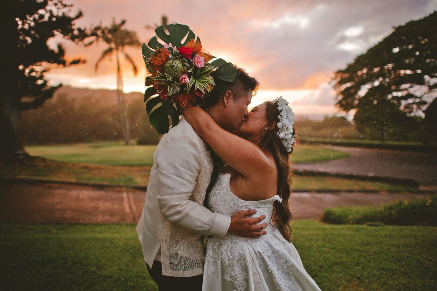 Couple sharing romantic kiss at sunset, groom wearing tropical leaf crown, lush Hawaiian garden.