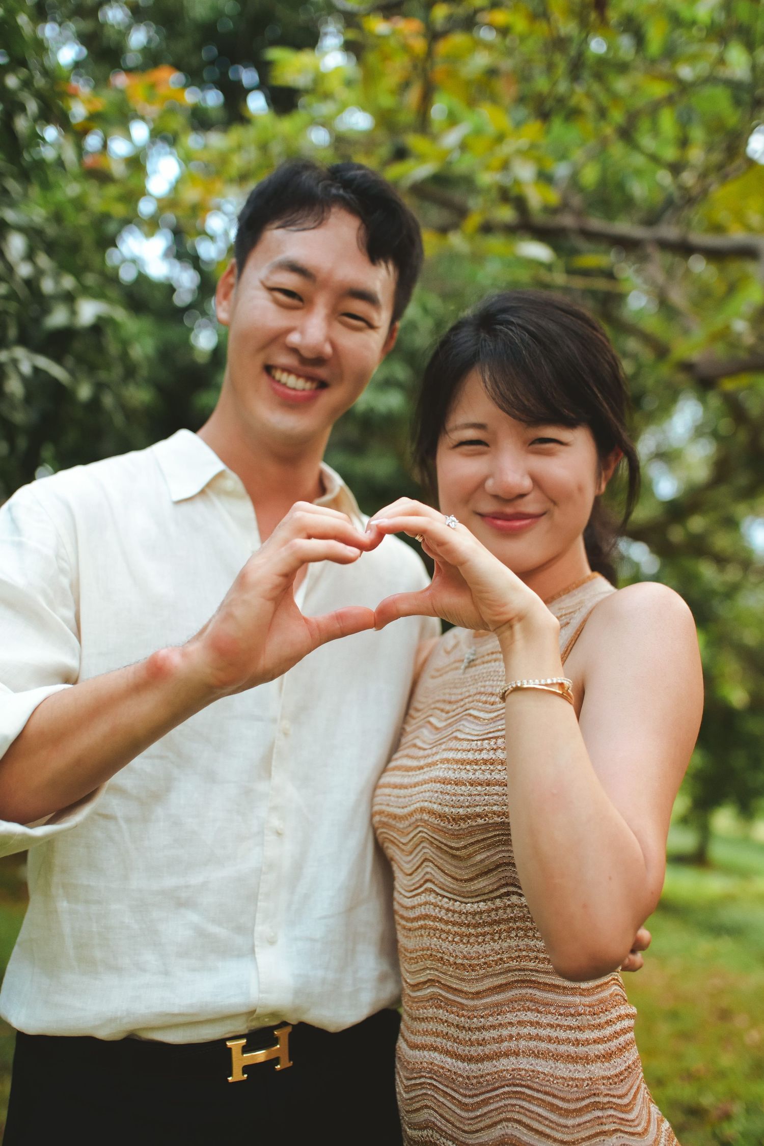 Happy Asian couple making a heart shape with their hands together outdoors in a green park setting.
