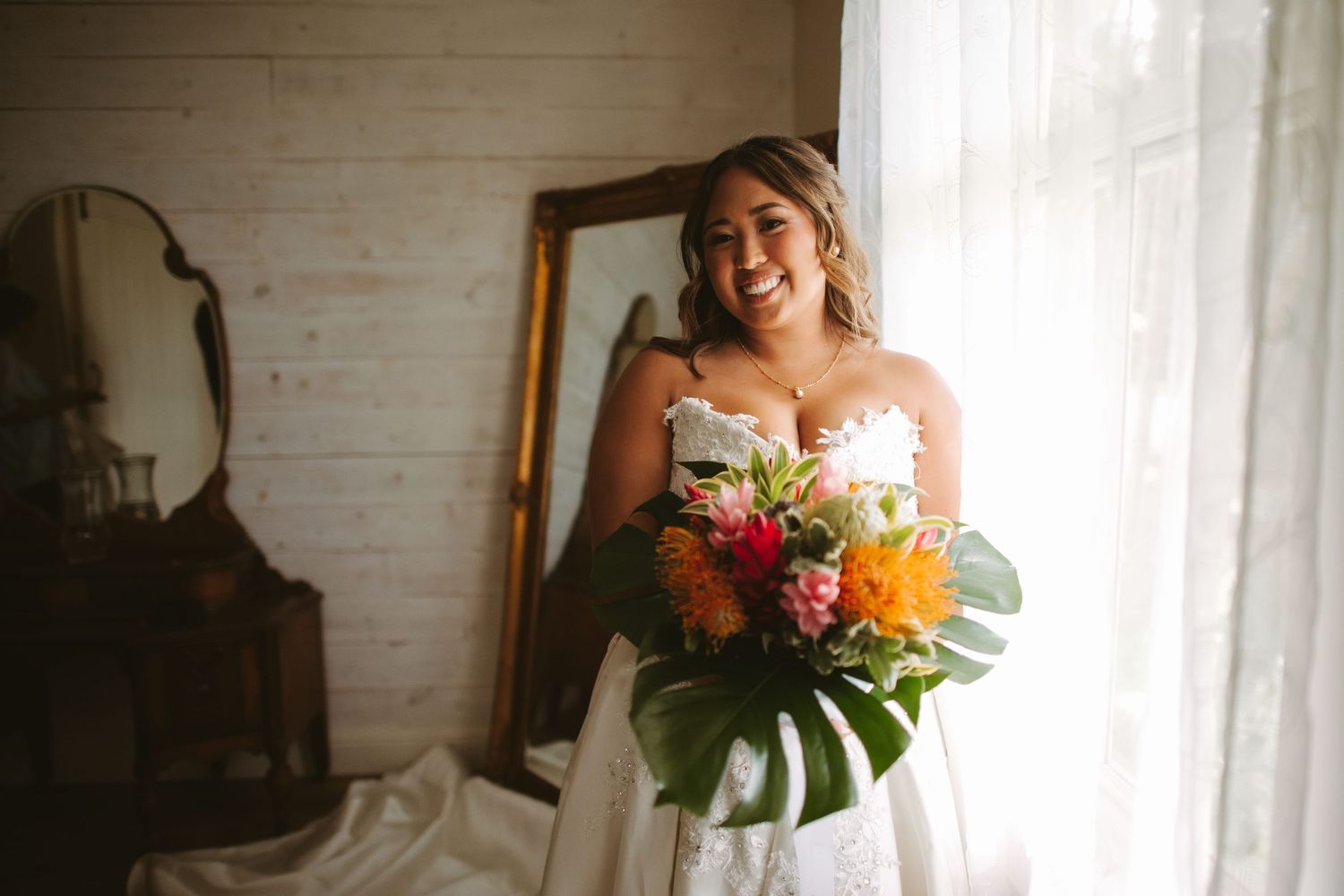Smiling bride in strapless wedding dress holding vibrant tropical bouquet with monstera leaves near window.