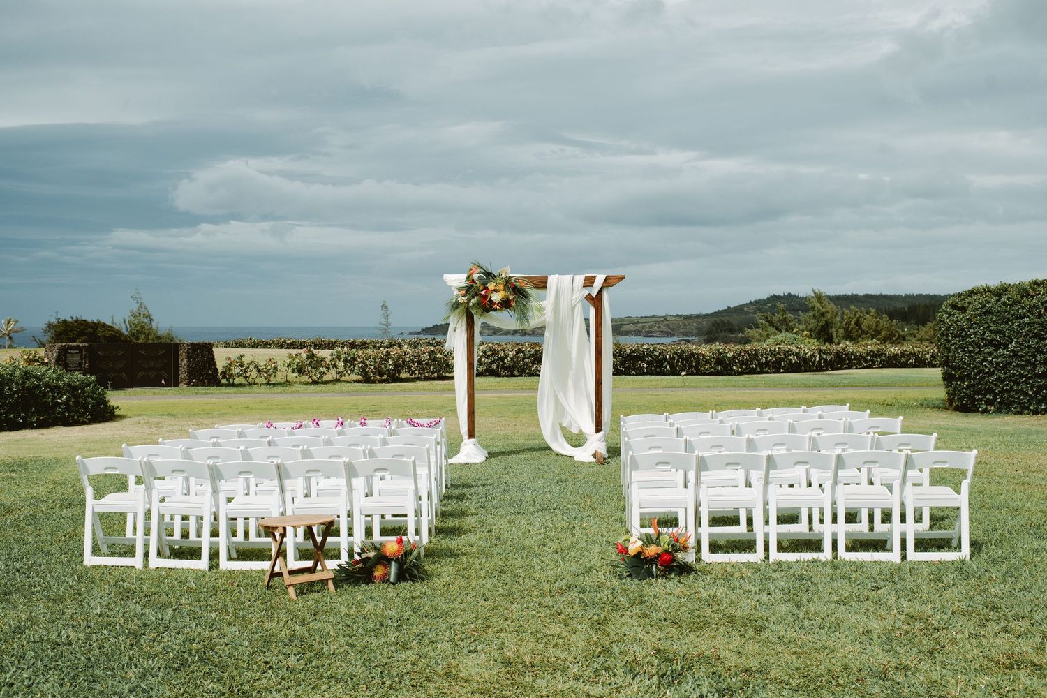 Outdoor wedding ceremony setup with white chairs, floral arch, and ocean views under cloudy skies.