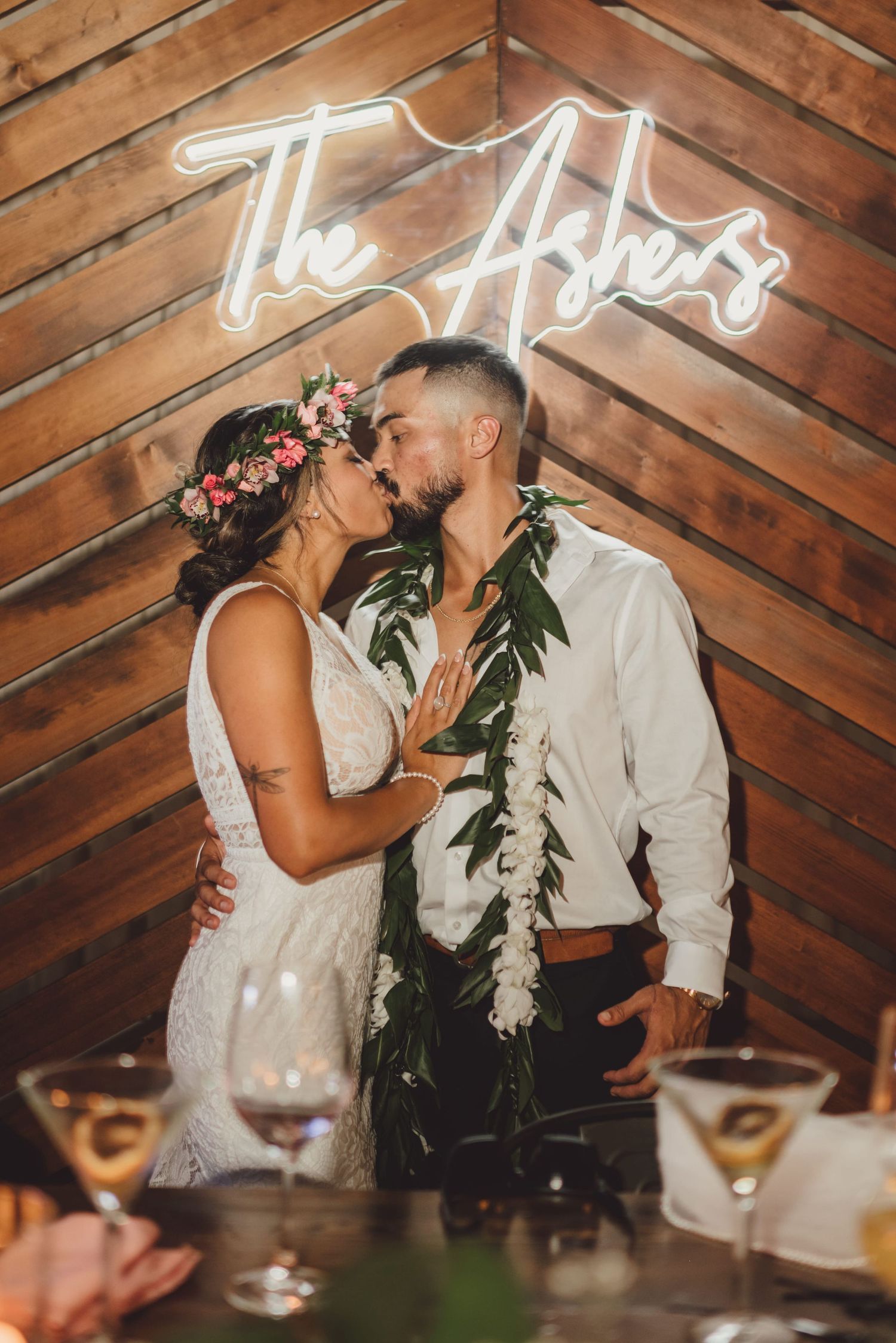 Newlyweds kissing under a neon sign reading The Ashers, groom wearing floral lei, bride in lace dress with flower crown.