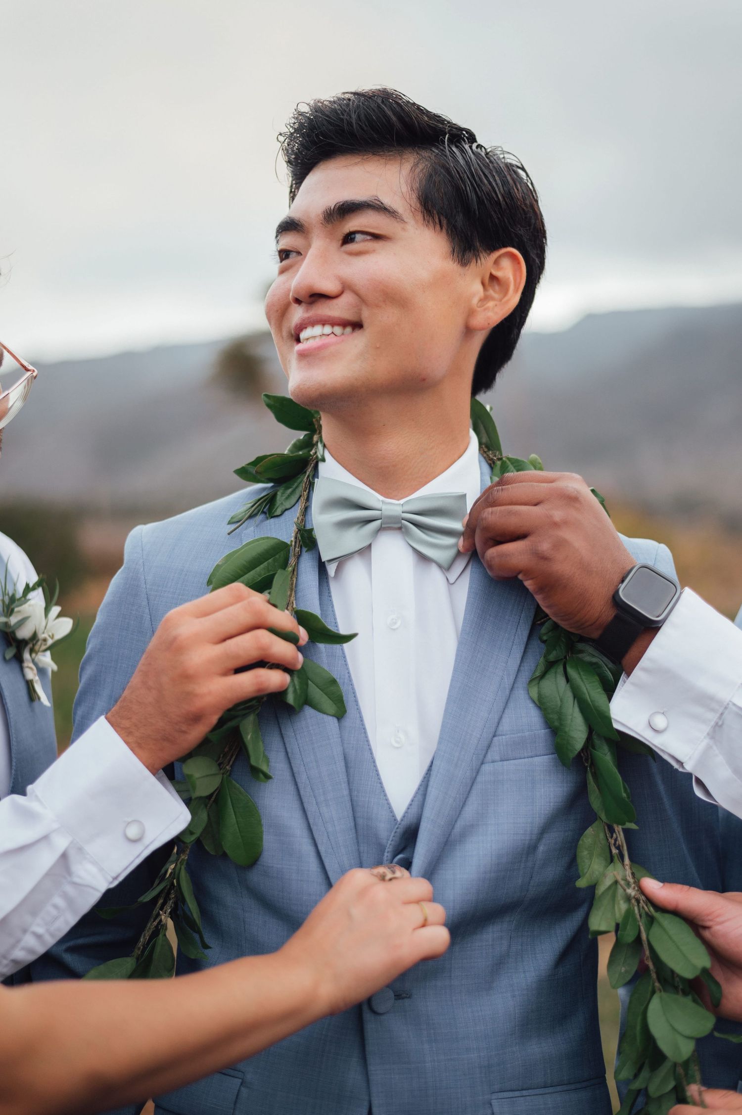 Smiling groom in light blue suit with green lei and sage bow tie being adjusted outdoors with mountains in background.