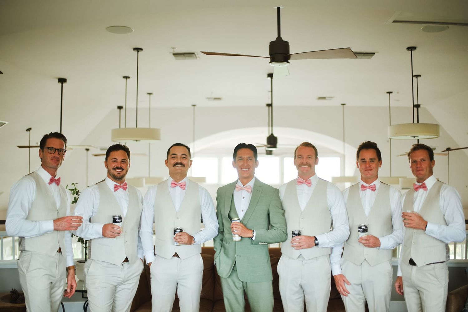 Groom in sage suit with groomsmen in light gray suits and pink bow ties posing indoors at a wedding venue.