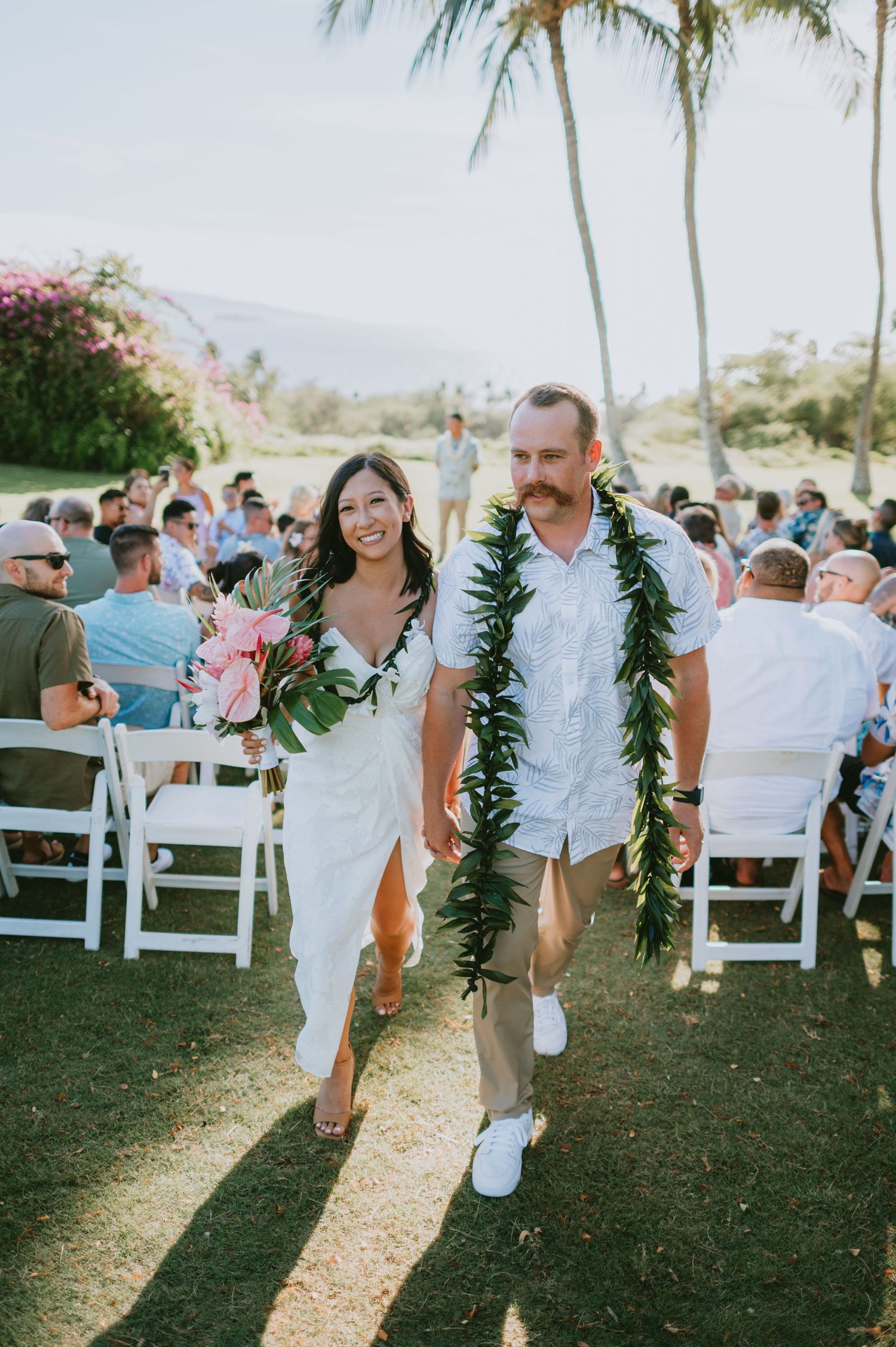 Couple walks down aisle in Hawaiian wedding, bride holds tropical bouquet, groom wears green lei, outdoor ceremony.