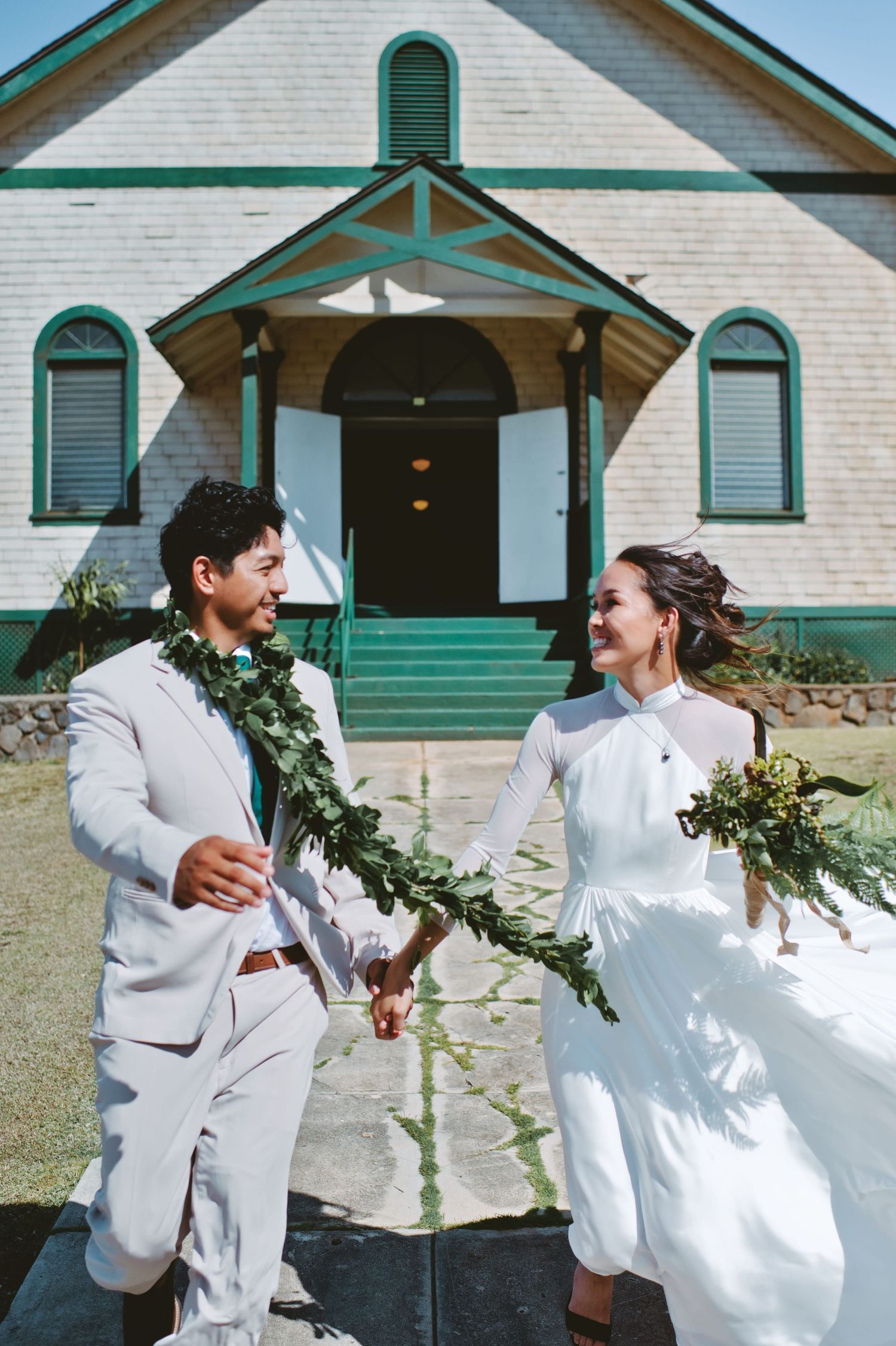 Bride and groom holding hands with Hawaiian lei garlands outside a charming green and white chapel on their wedding day.