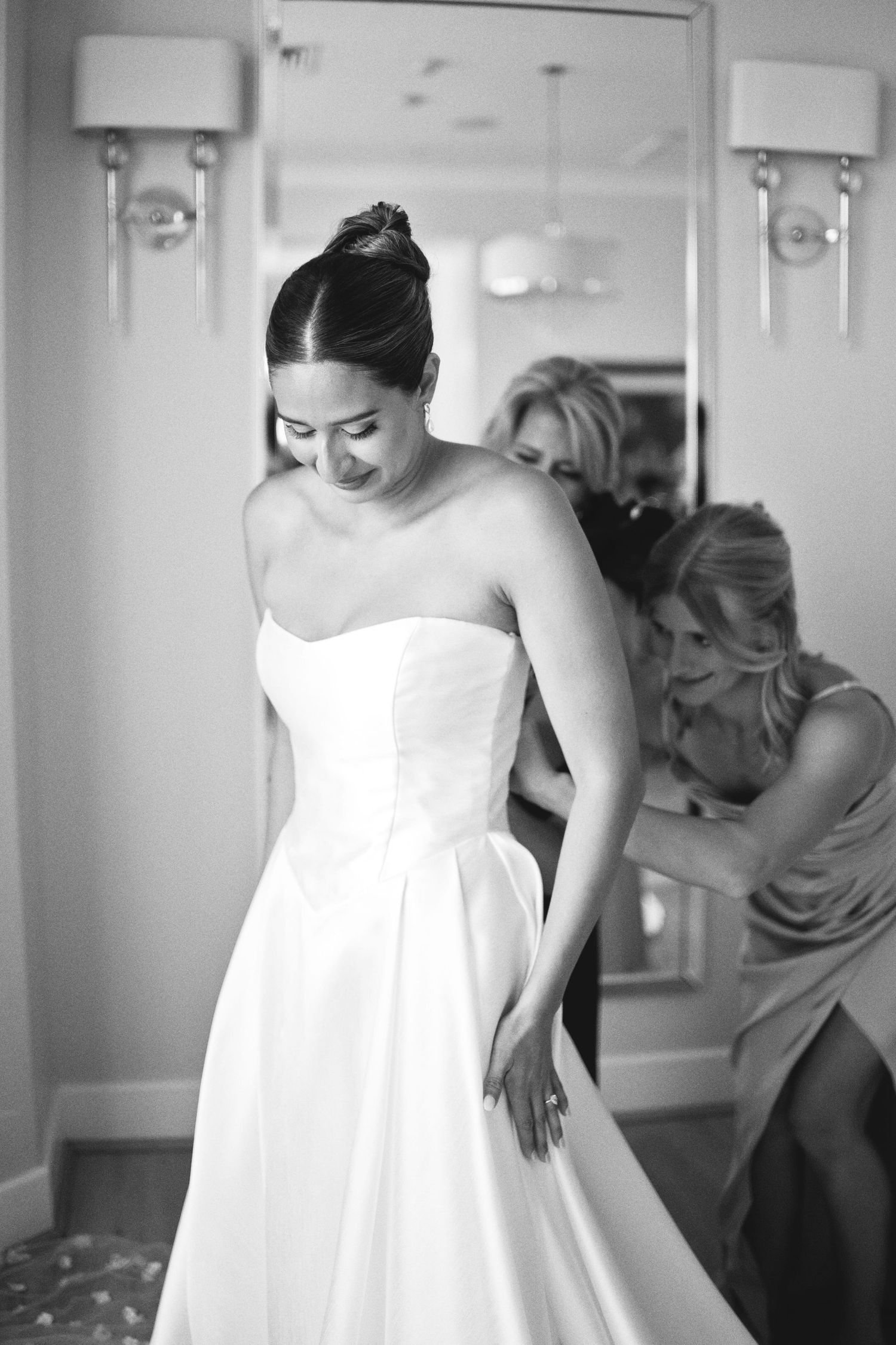 Black and white photo of a bride in a strapless gown smiling as bridesmaid helps fasten her wedding dress.