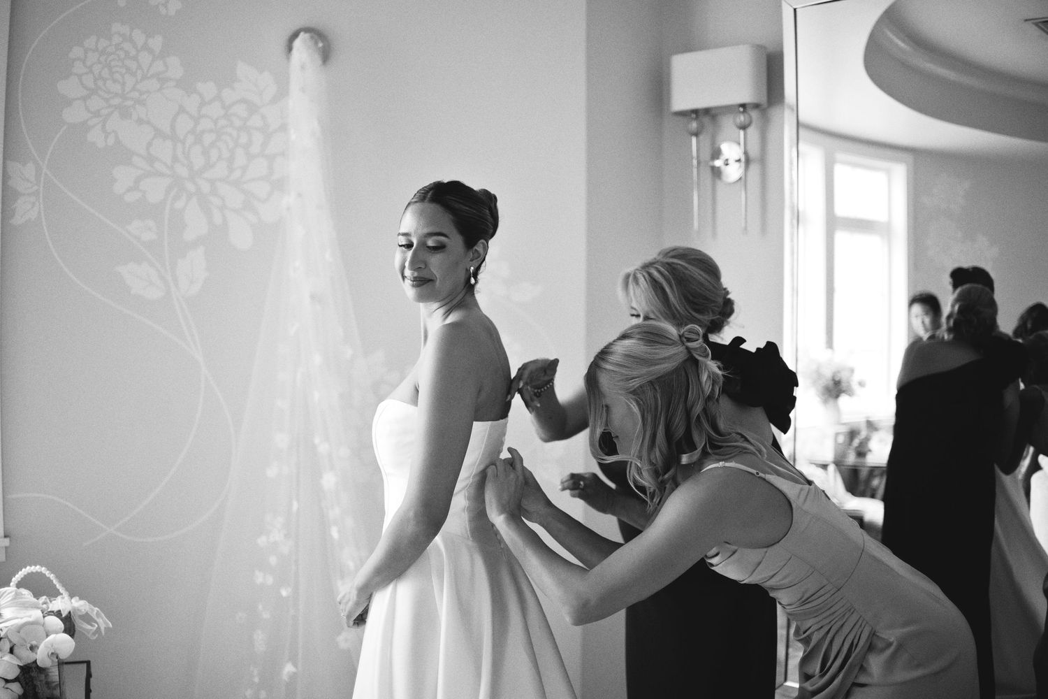 Black and white photo of a bride in a white gown and veil being helped by bridesmaids before her wedding ceremony.