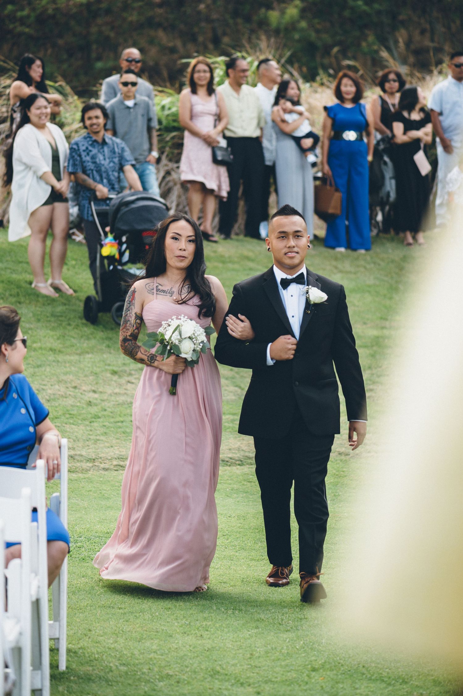 Wedding party members walking down the aisle in formal attire on a grassy lawn.