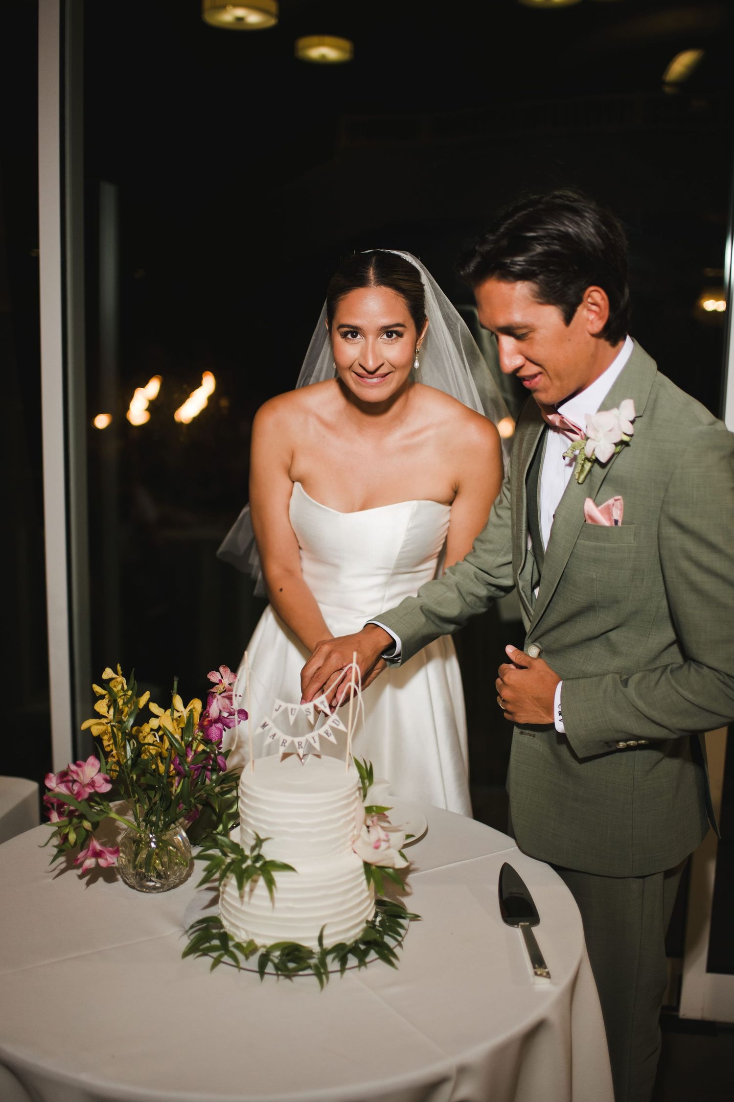 Bride and groom cutting a white tiered wedding cake together, bride in strapless gown with veil, groom in green suit.