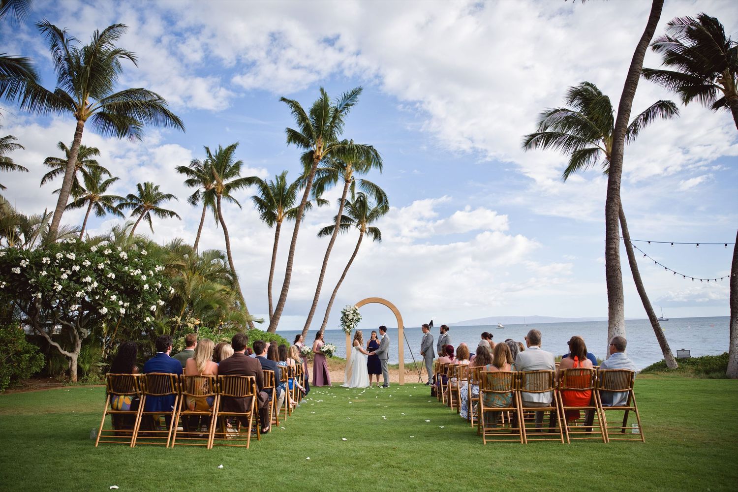 Outdoor beach wedding ceremony on lush green lawn with palm trees and ocean backdrop in Hawaii.