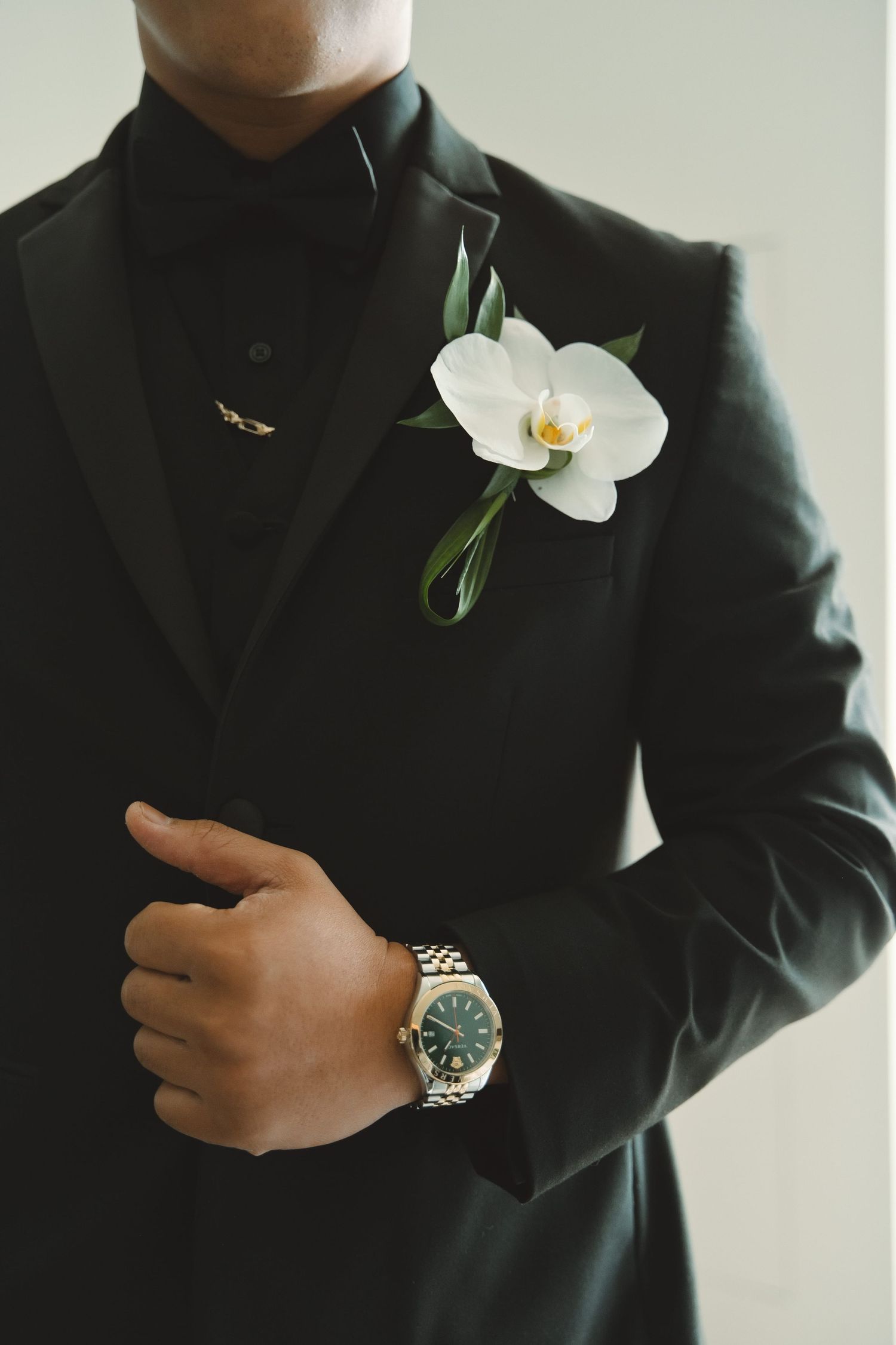 Groom in black tuxedo with white orchid boutonniere and gold Rolex watch, adjusting jacket lapel.