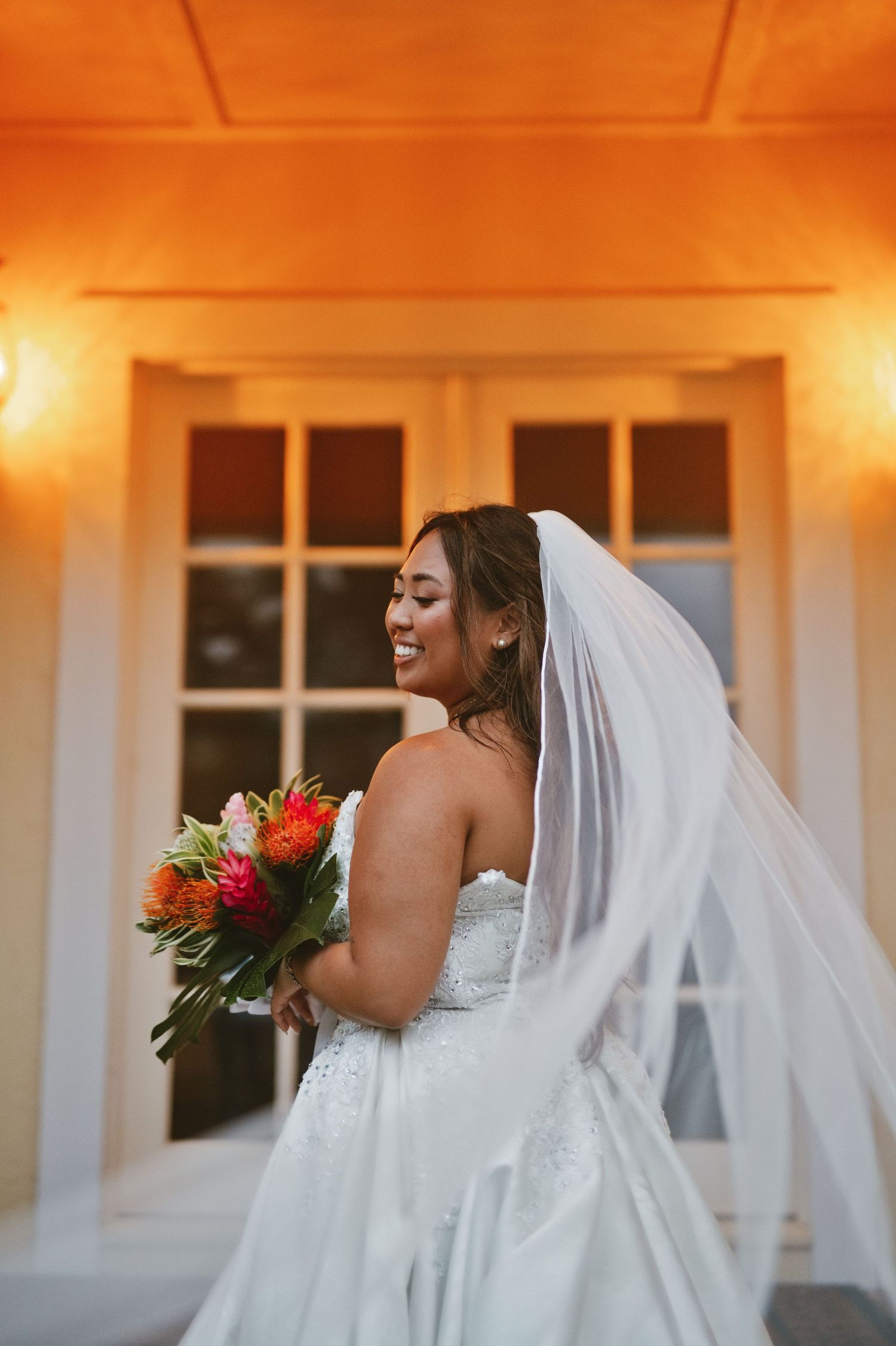 Smiling bride in strapless lace gown and flowing veil holds vibrant tropical bouquet outdoors in warm golden light.