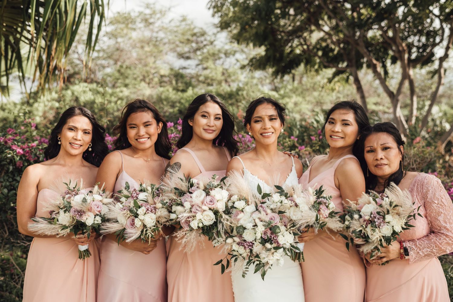 Bride in white gown with five bridesmaids in blush pink dresses holding floral bouquets outdoors.