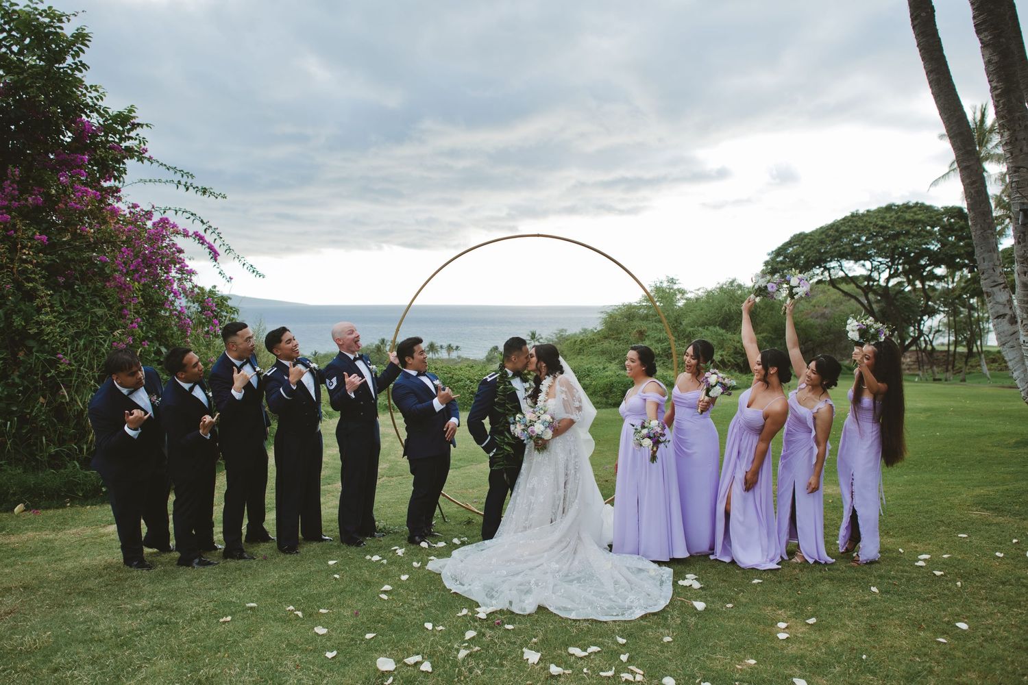 Wedding party poses under circular arch by ocean, bridesmaids in lavender gowns, groomsmen in dark suits, bride and groom kissing.
