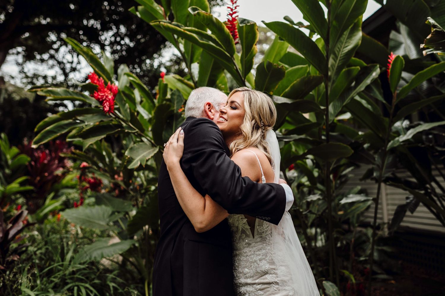Bride and older man share emotional hug surrounded by lush tropical greenery and red flowers at outdoor wedding.