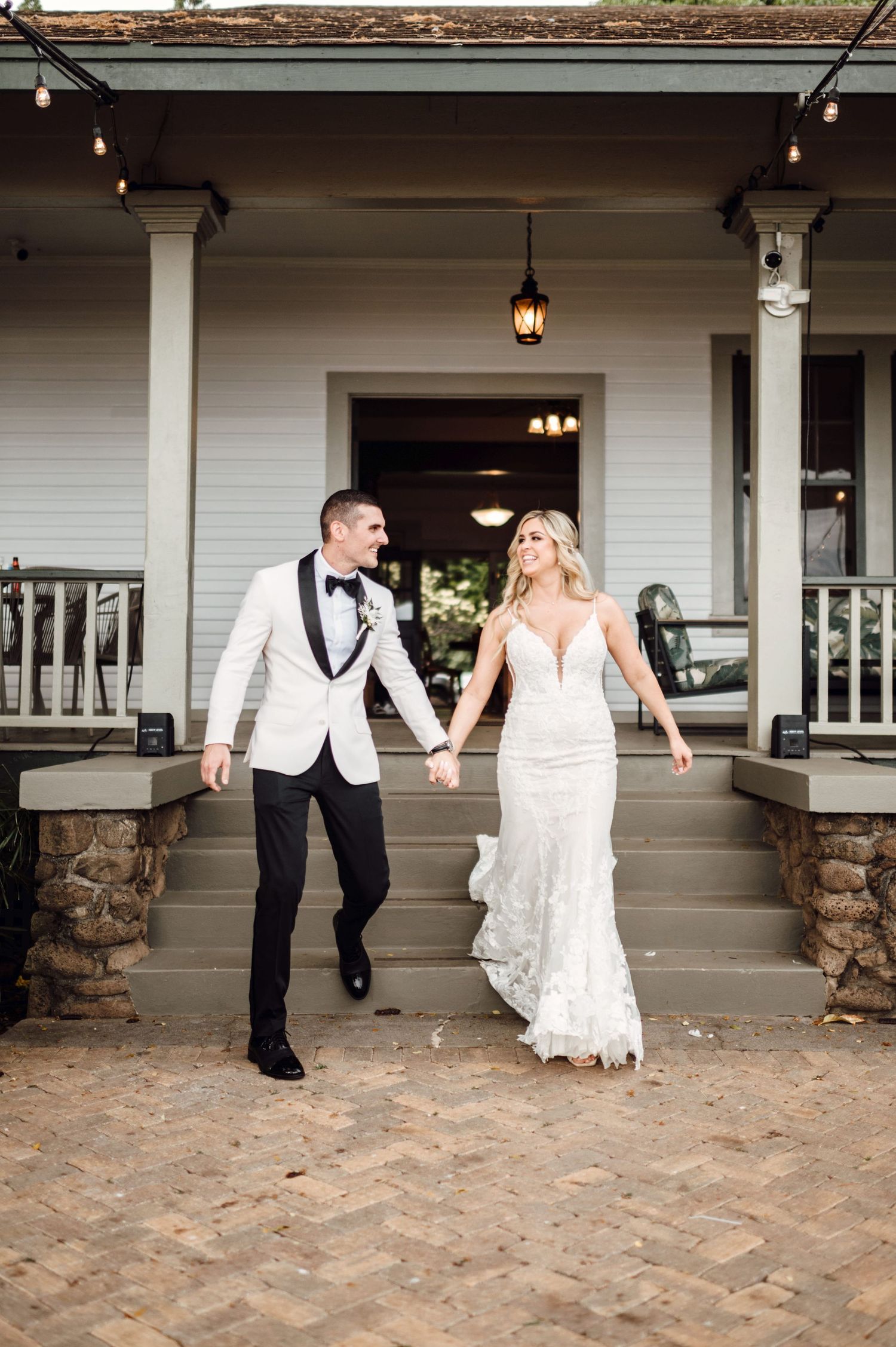 Bride in lace gown and groom in white tuxedo jacket hold hands walking down brick steps at elegant wedding venue.