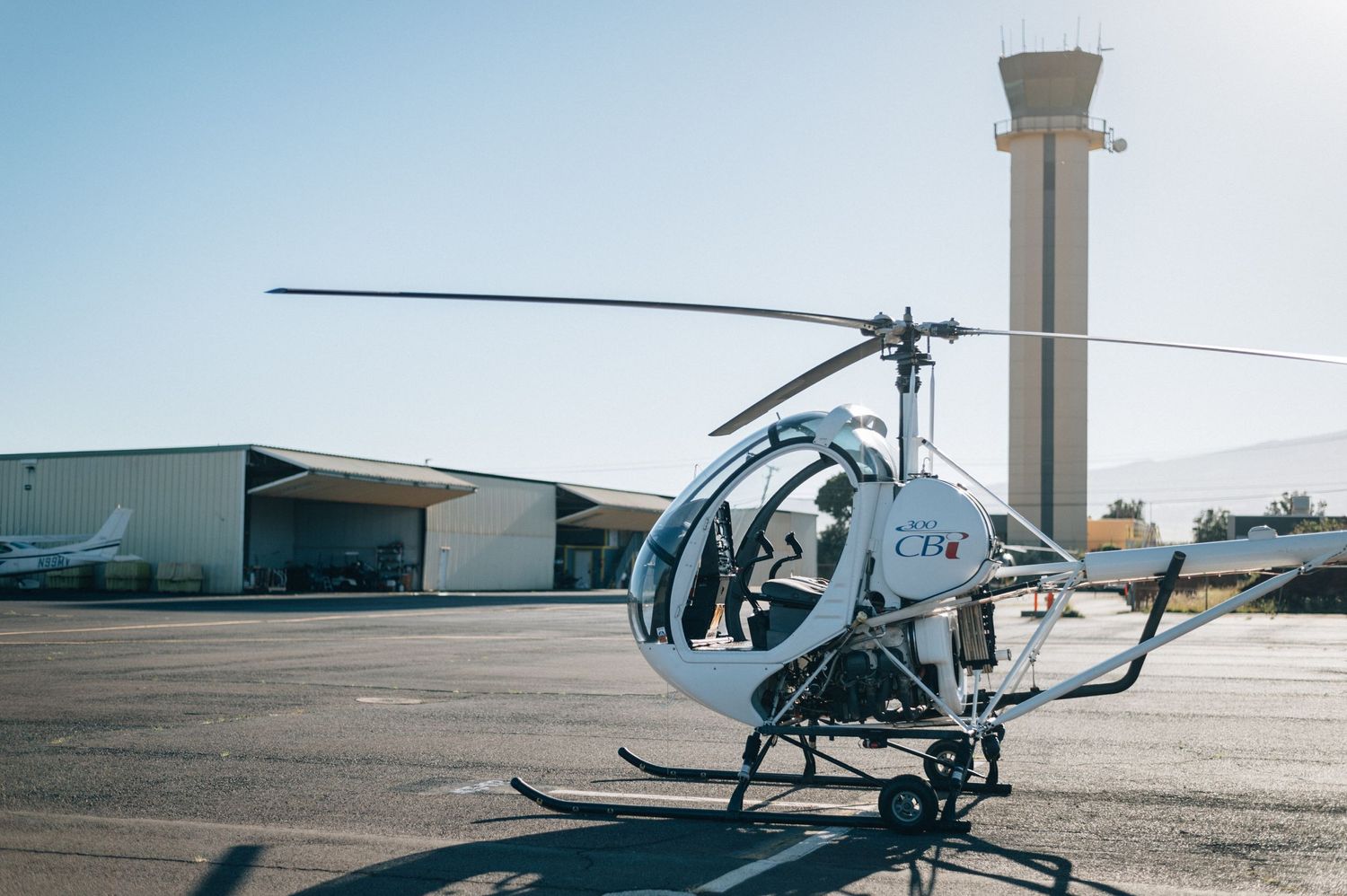 Small white helicopter parked near airport control tower under clear sky.